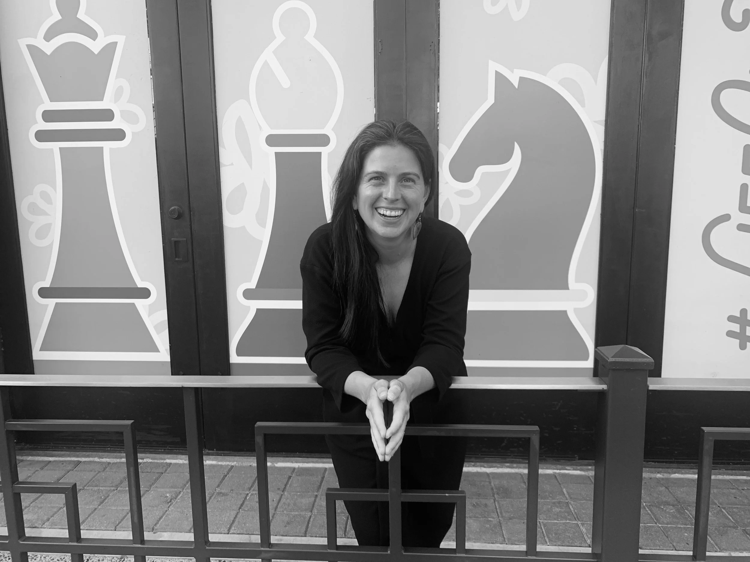 Joanna, smiling, leaning on a metal fence in front of large chess piece graphics on a wall.