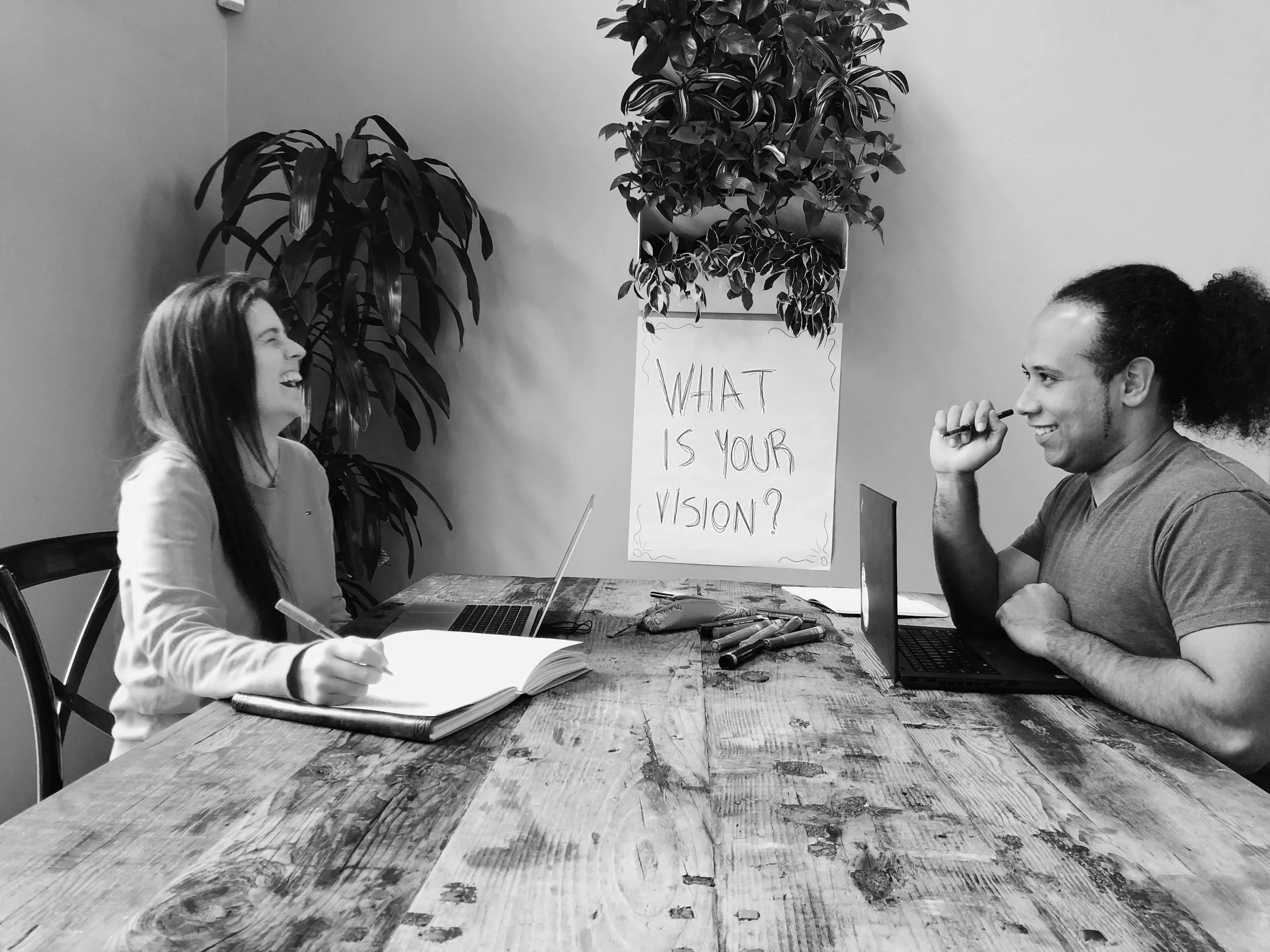 Two people sitting across a wooden table, having a conversation and smiling. A plant and a poster with the handwritten question 'What is your vision?' are visible in the background.