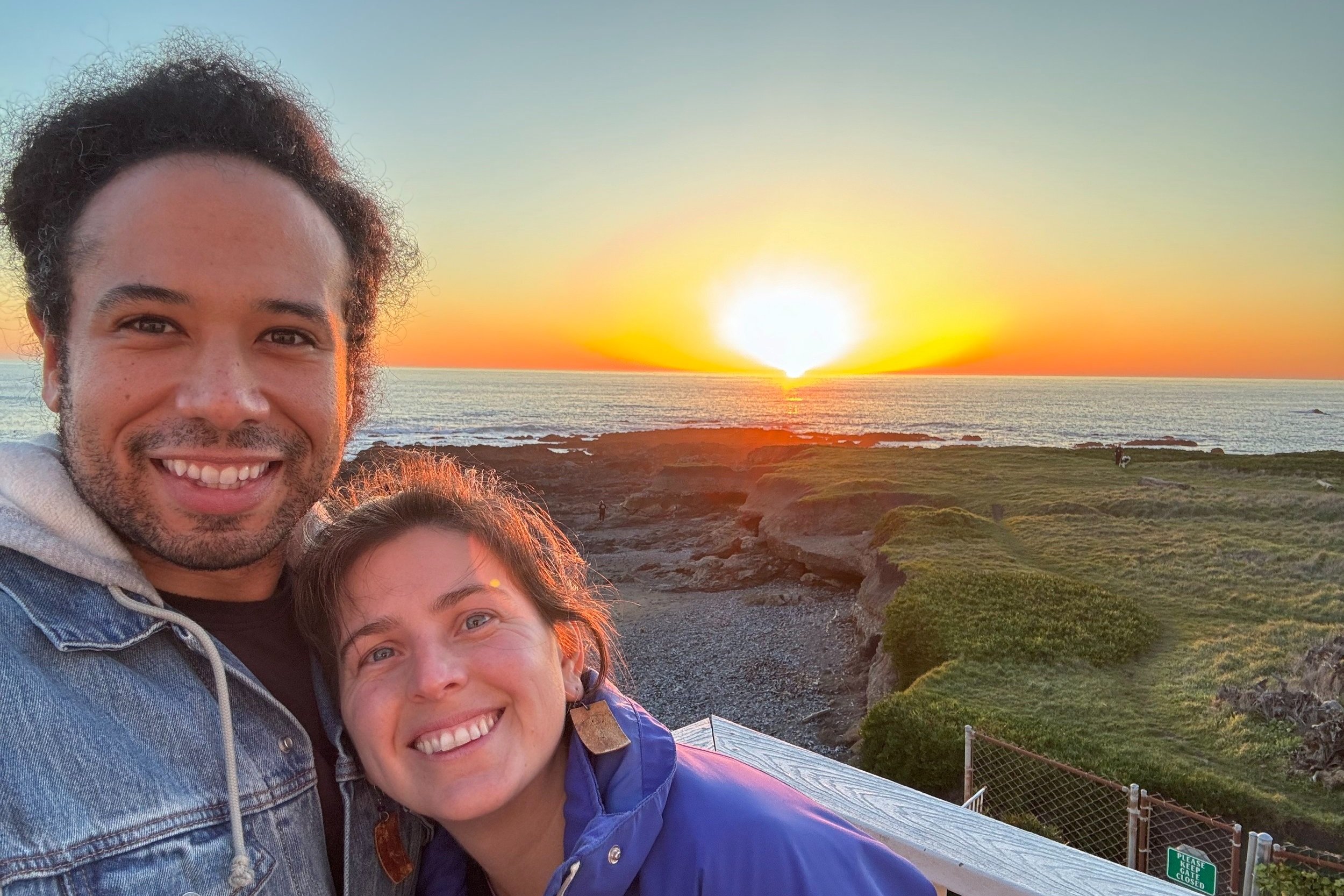 The image captures two people standing on a rocky outcrop overlooking the ocean during sunset. The sun’s warm glow bathes the scene in a golden hue, casting long shadows on the ground. The water stretches out before them, its surface shimmering.