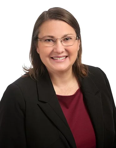 Professional woman with glasses, wearing a black blazer and maroon top, smiling against a plain white background.