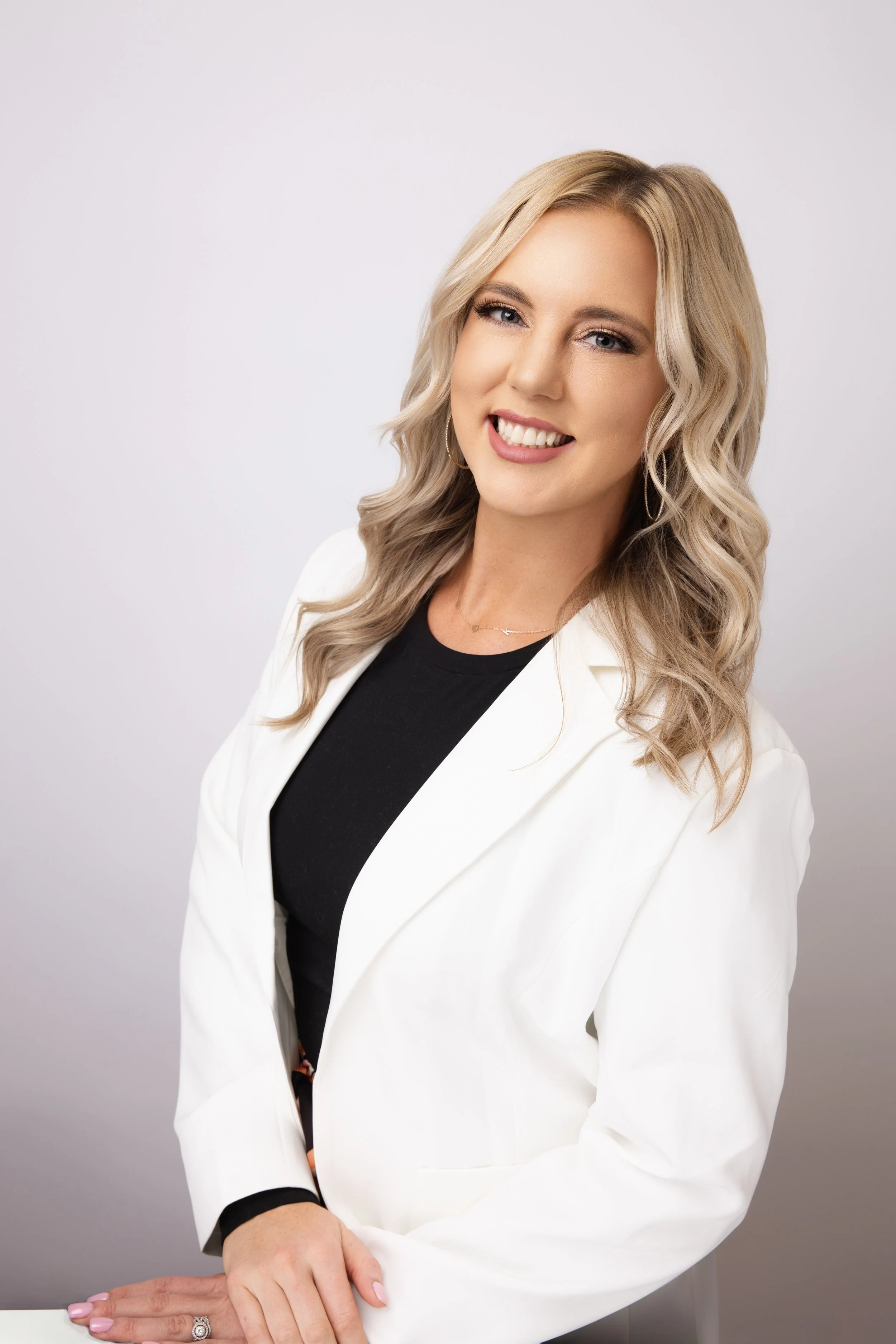 A young woman with blonde wavy hair, wearing a white blazer over a black shirt, smiling and looking at the camera.