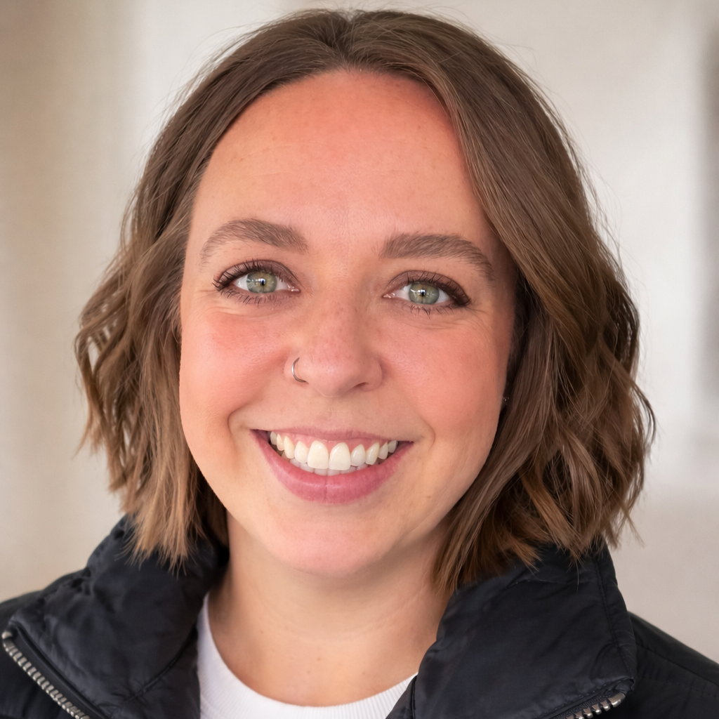 A smiling woman with short, wavy brown hair, light skin, and green eyes, wearing a black jacket over a white shirt.