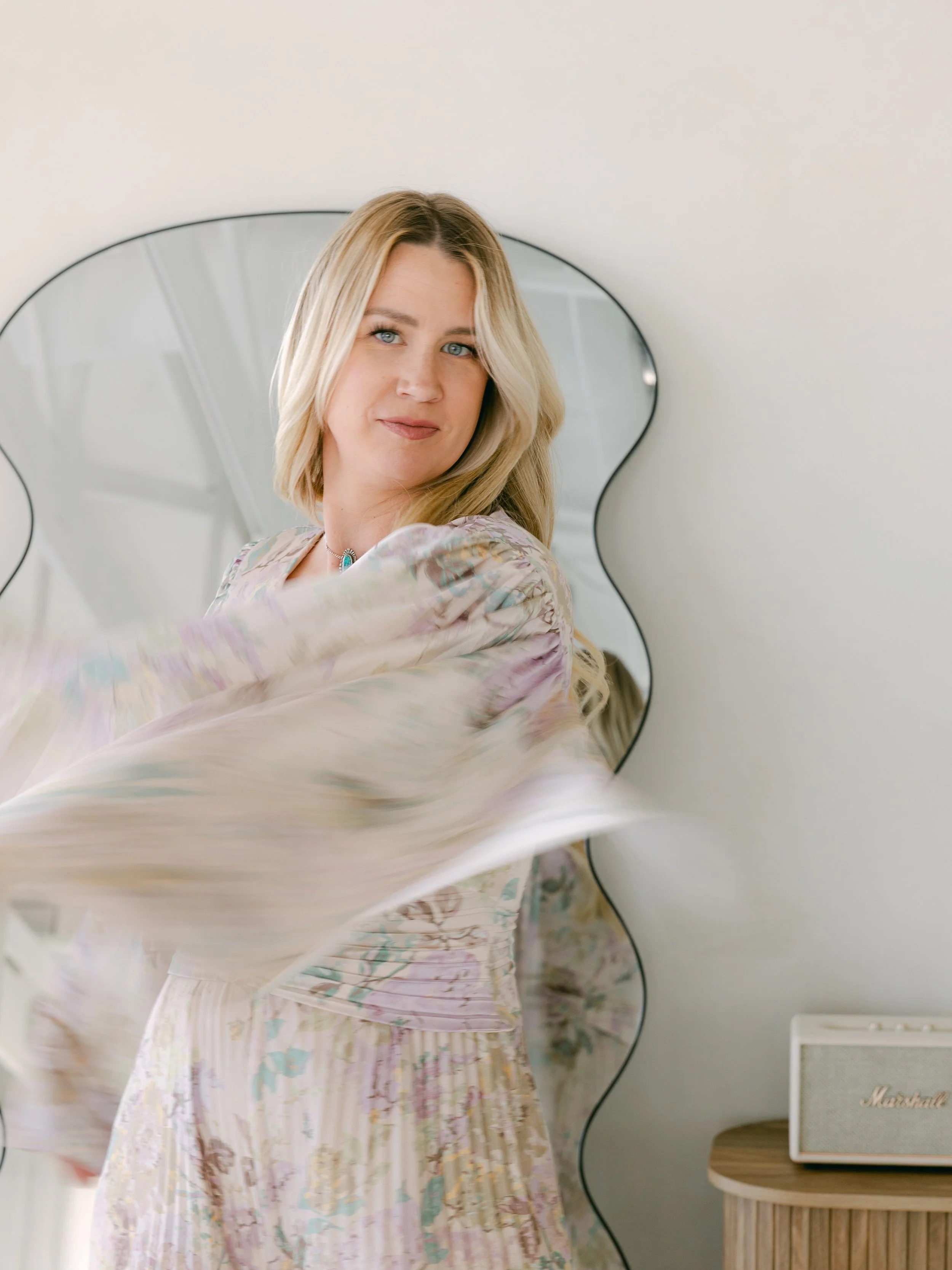 A blonde woman with shoulder-length hair wearing a light-colored, flowy, floral-patterned dress, standing in a bright room with a decorative mirror and a Marshall speaker in the background.