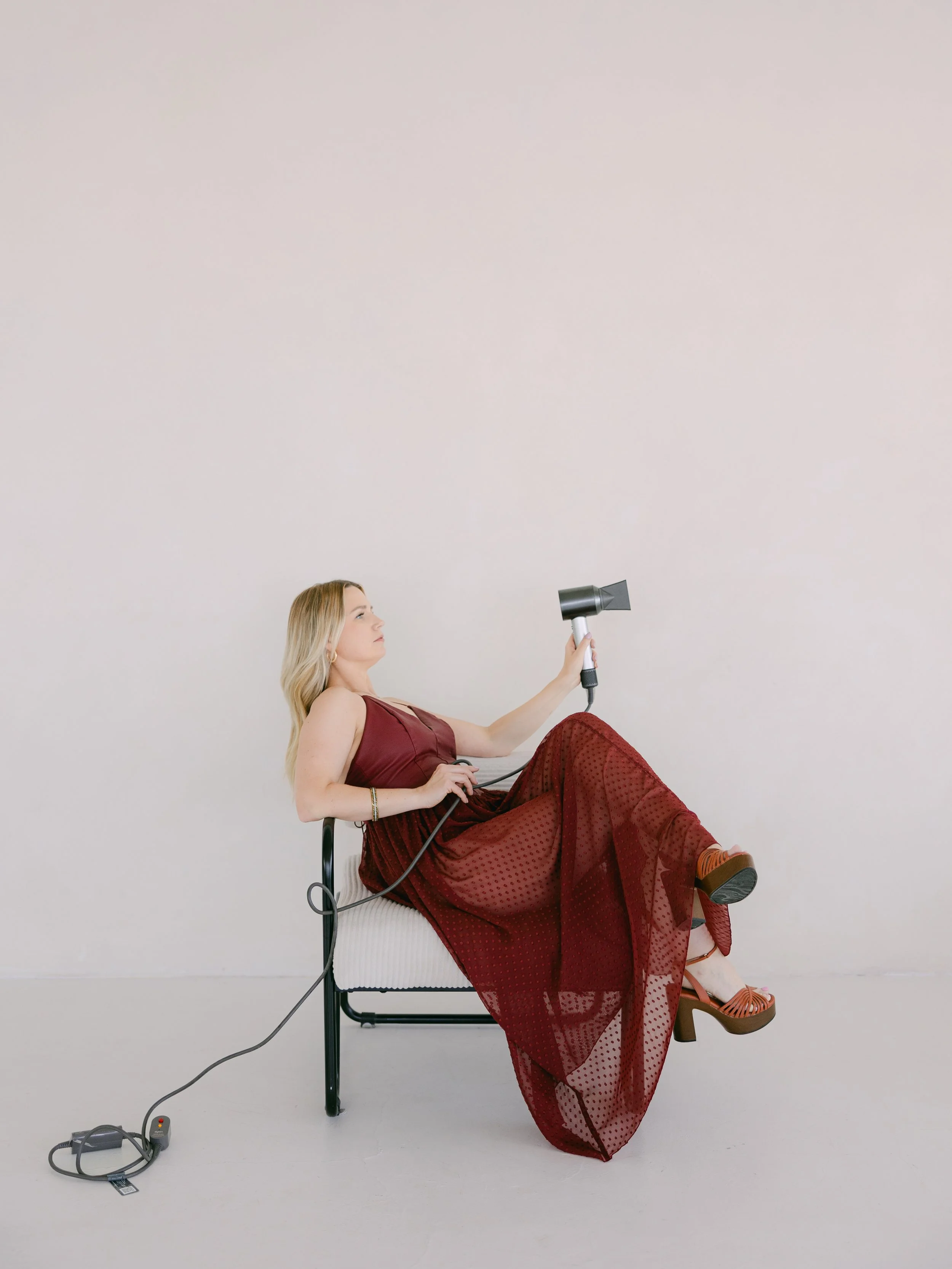 A woman with long blonde hair wearing a red dress sitting on a black metal bench against a plain white wall. She is holding a black hairdryer in her right hand and a dog clipper in her left hand, with an electrical cord connected to a power strip on