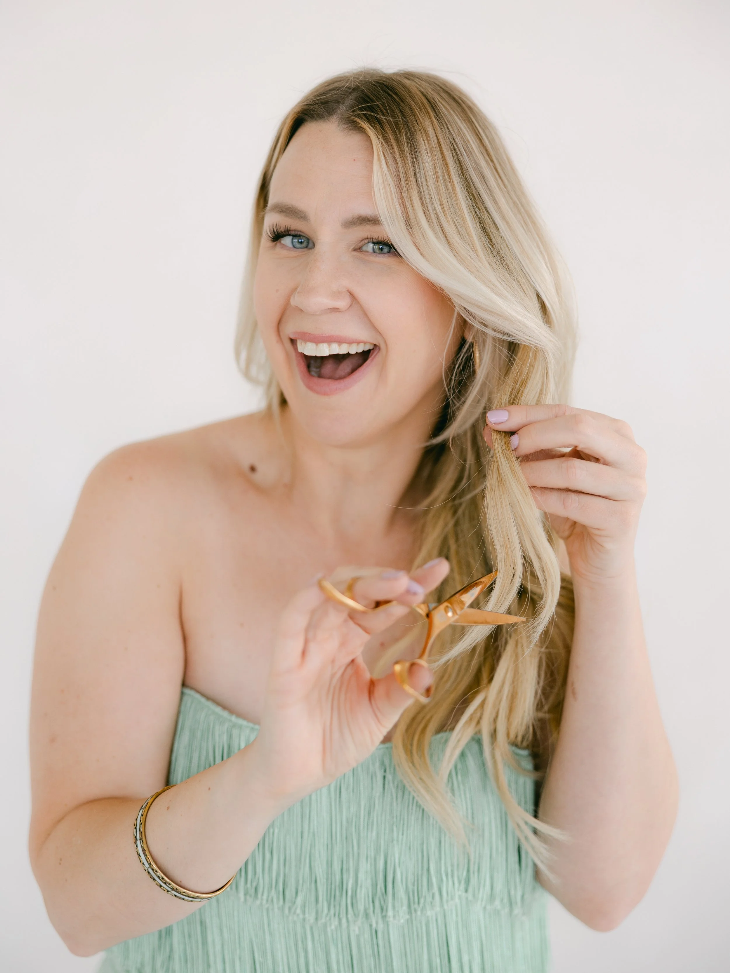 A woman with blonde hair, blue eyes, and a towel or dress, smiling while holding a pair of gold scissors and trimming her hair.