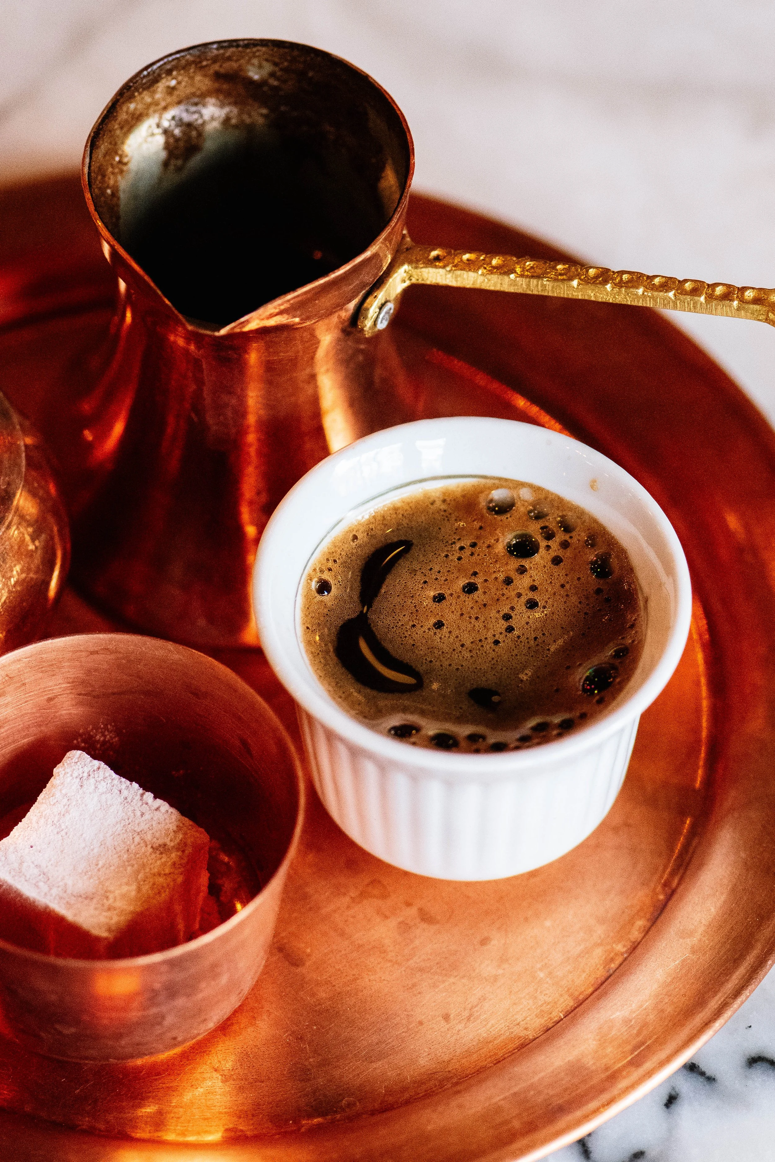 A cup of black coffee, a sugar cube in a small copper container, and a copper iced coffee pot on a copper tray.