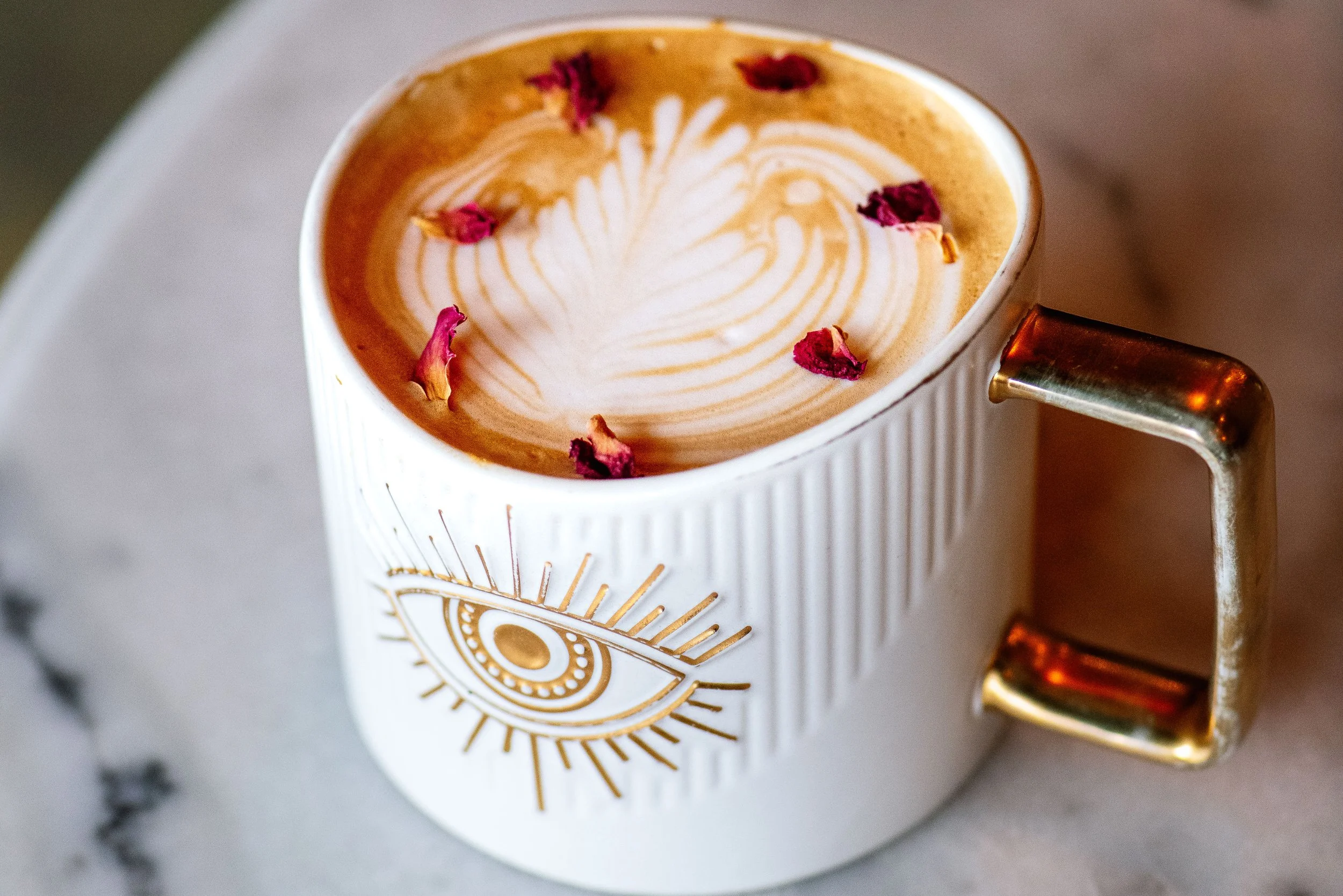 Close-up of a white ceramic mug with a gold eye design on the front, filled with latte with latte art and dried rose petals on top, sitting on a marble surface.