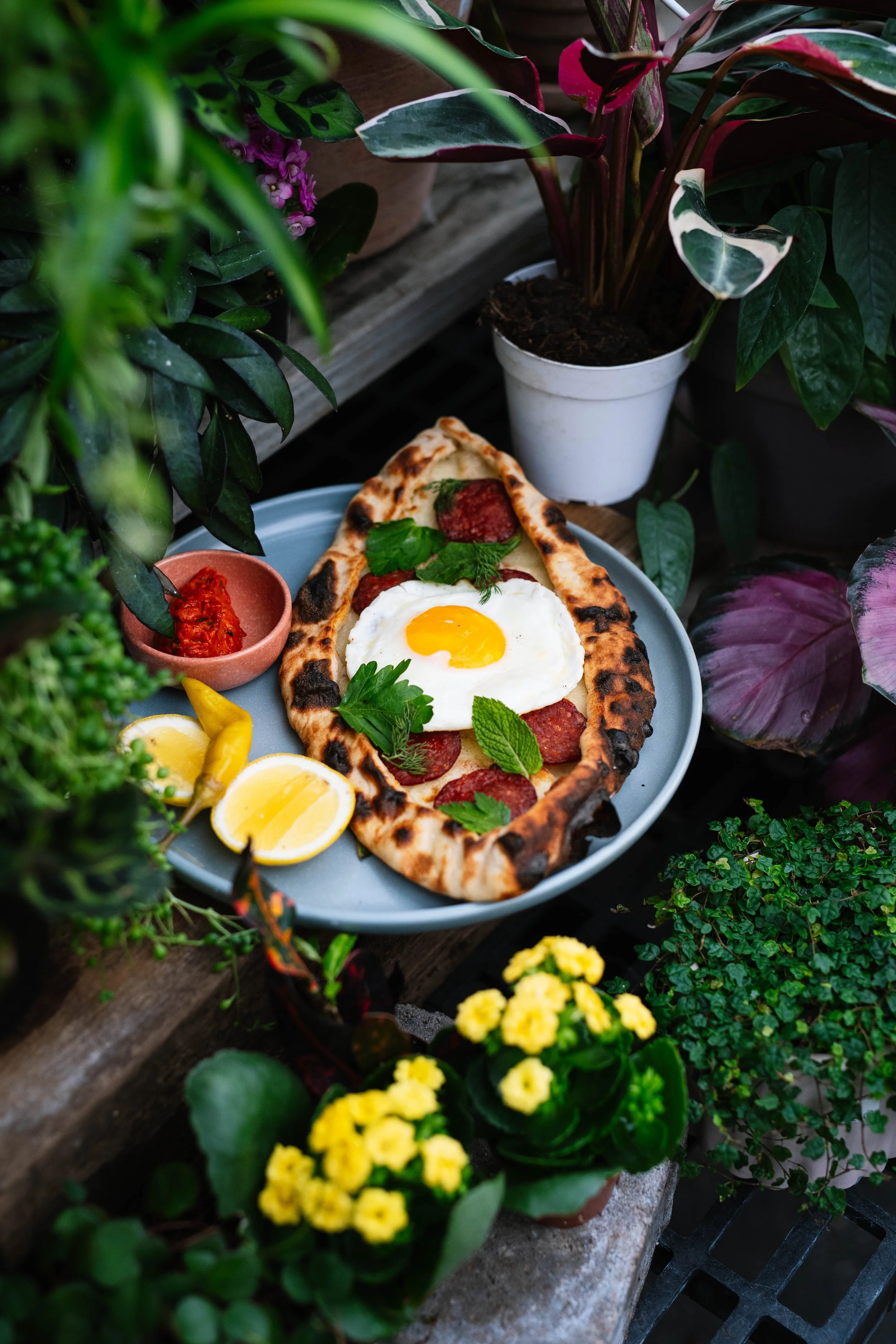 A plate with pepperoni pizza topped with a fried egg, garnished with parsley and mint, served with lemon wedges and a small bowl of red sauce, amidst potted plants and greenery.