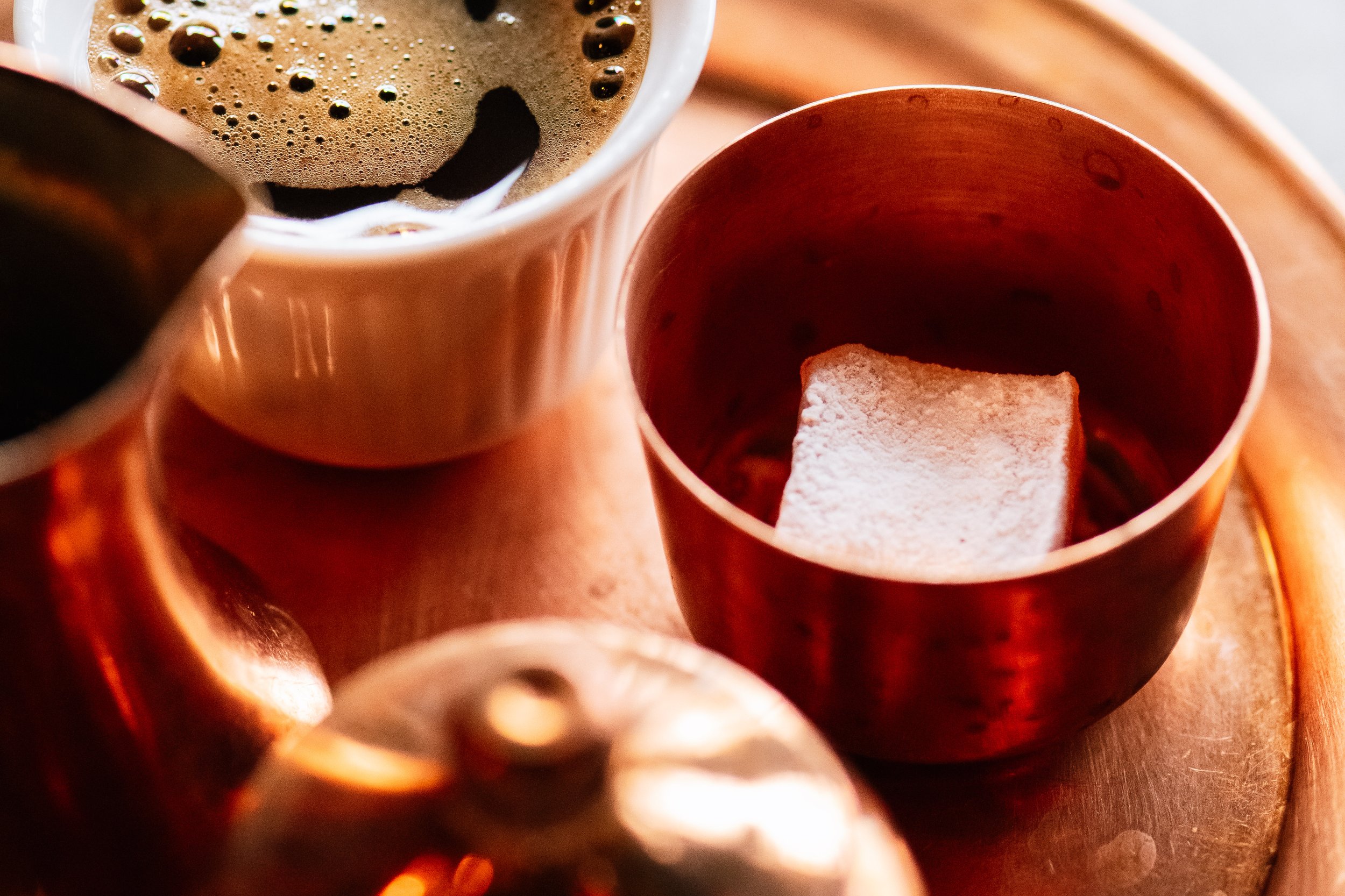 A copper cup containing a sugar cube, a white cup filled with coffee, and part of a teapot on a wooden tray.