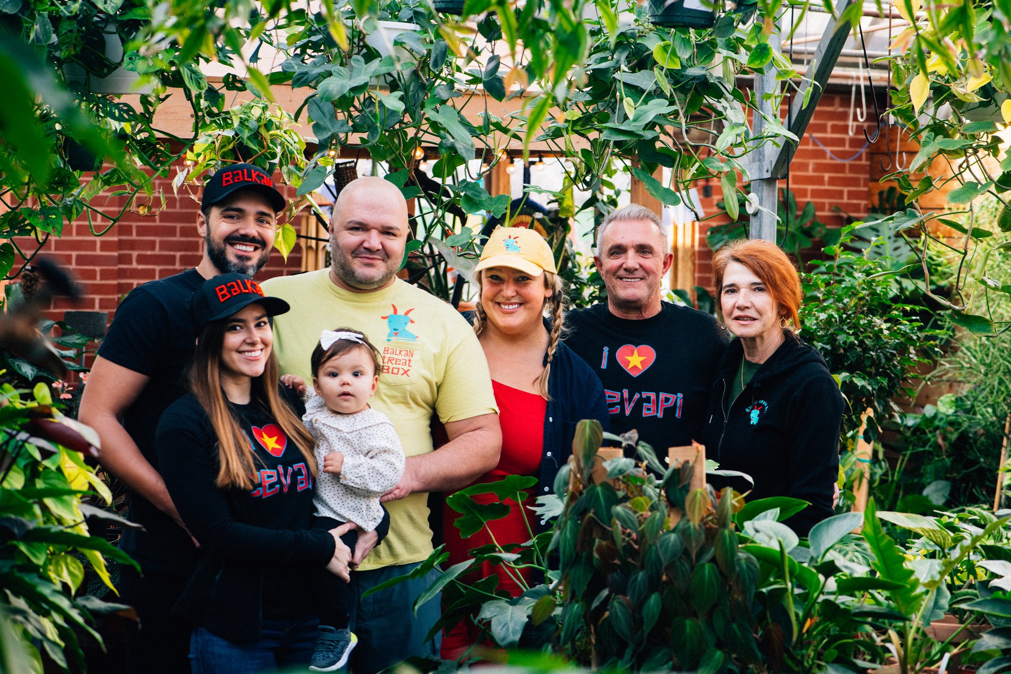 Group of six people, including a young girl and a baby, standing among lush green plants in a garden with a brick building in the background, smiling at the camera.