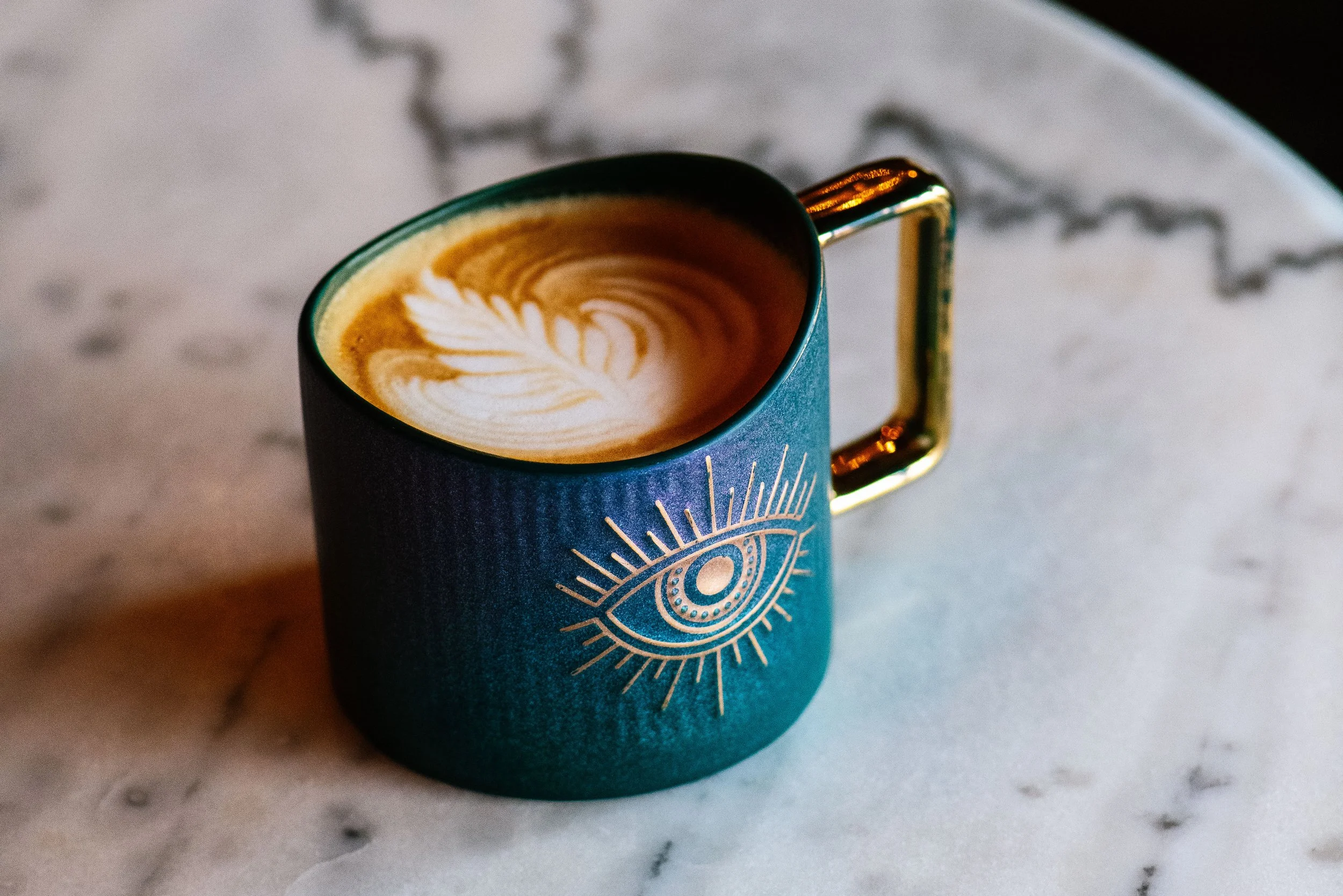 A blue coffee mug with a gold handle, featuring an eye design with radiating lines, sitting on a white marble surface. The mug contains a latte with foam art resembling a leaf.