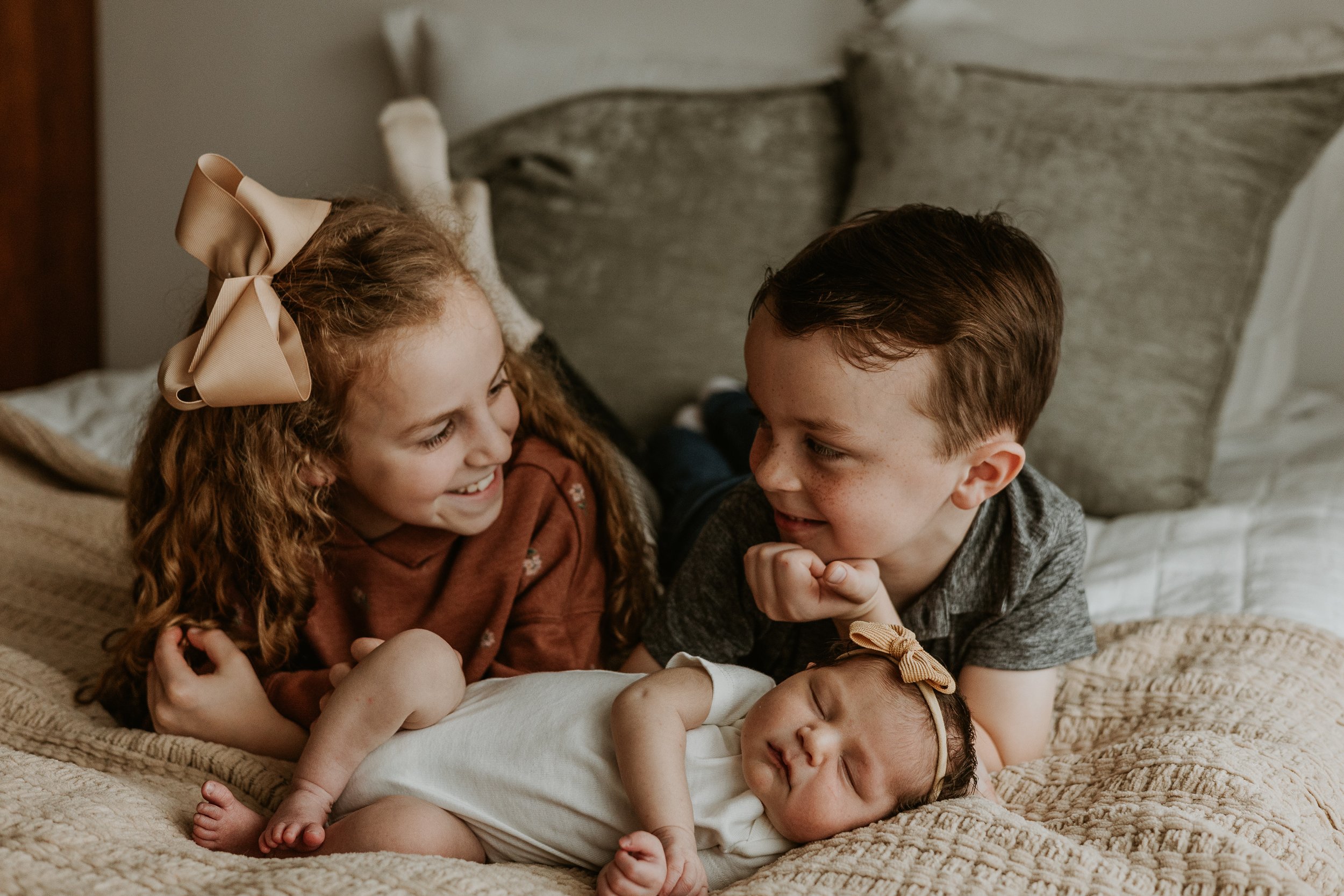 A big brother and big sister smile at each other while their baby sister sleeps peacefull in front of them.