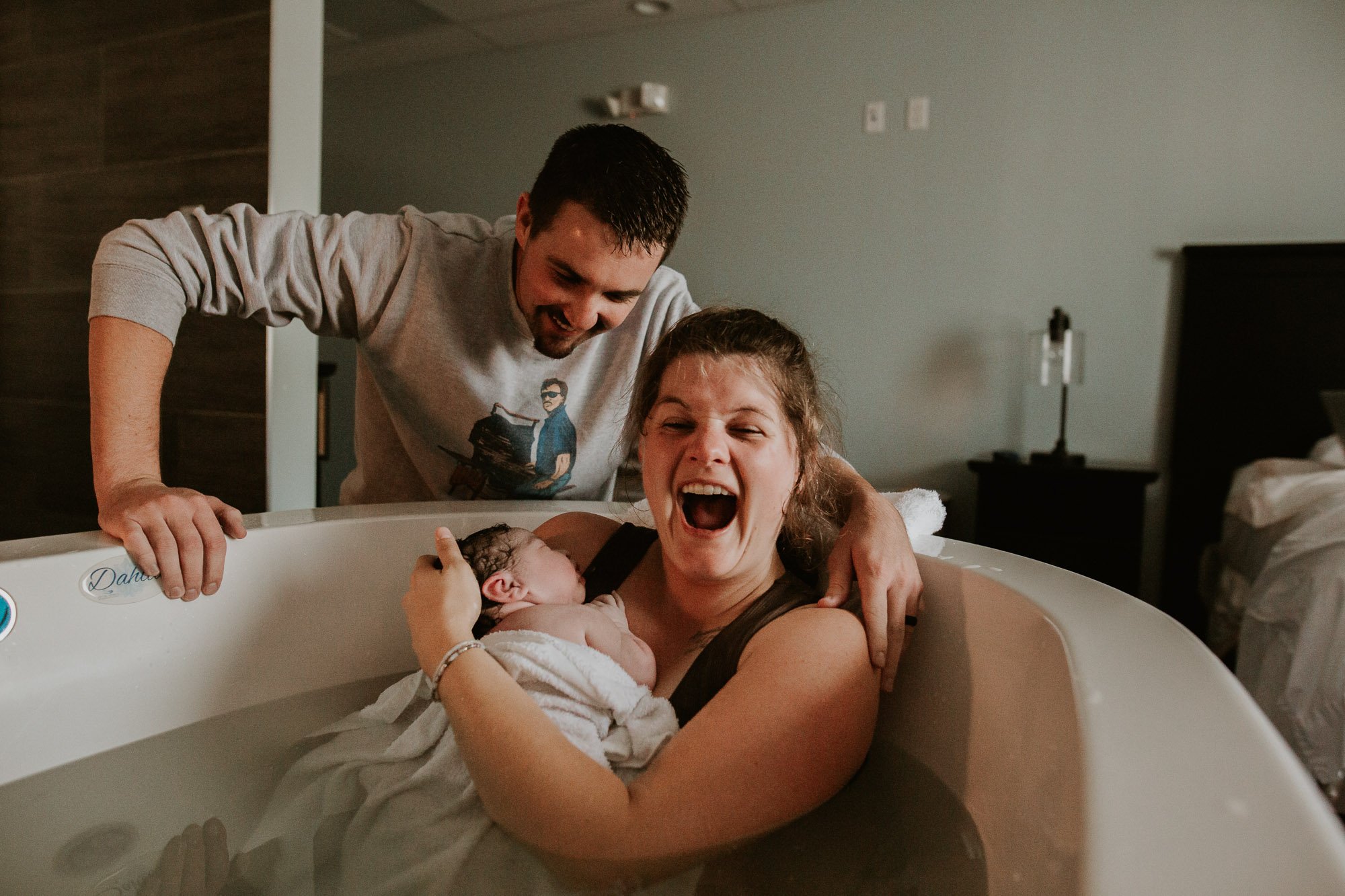 A mom holds her newborn baby in a birth tub and smiles elatedly at the camera after her birth.