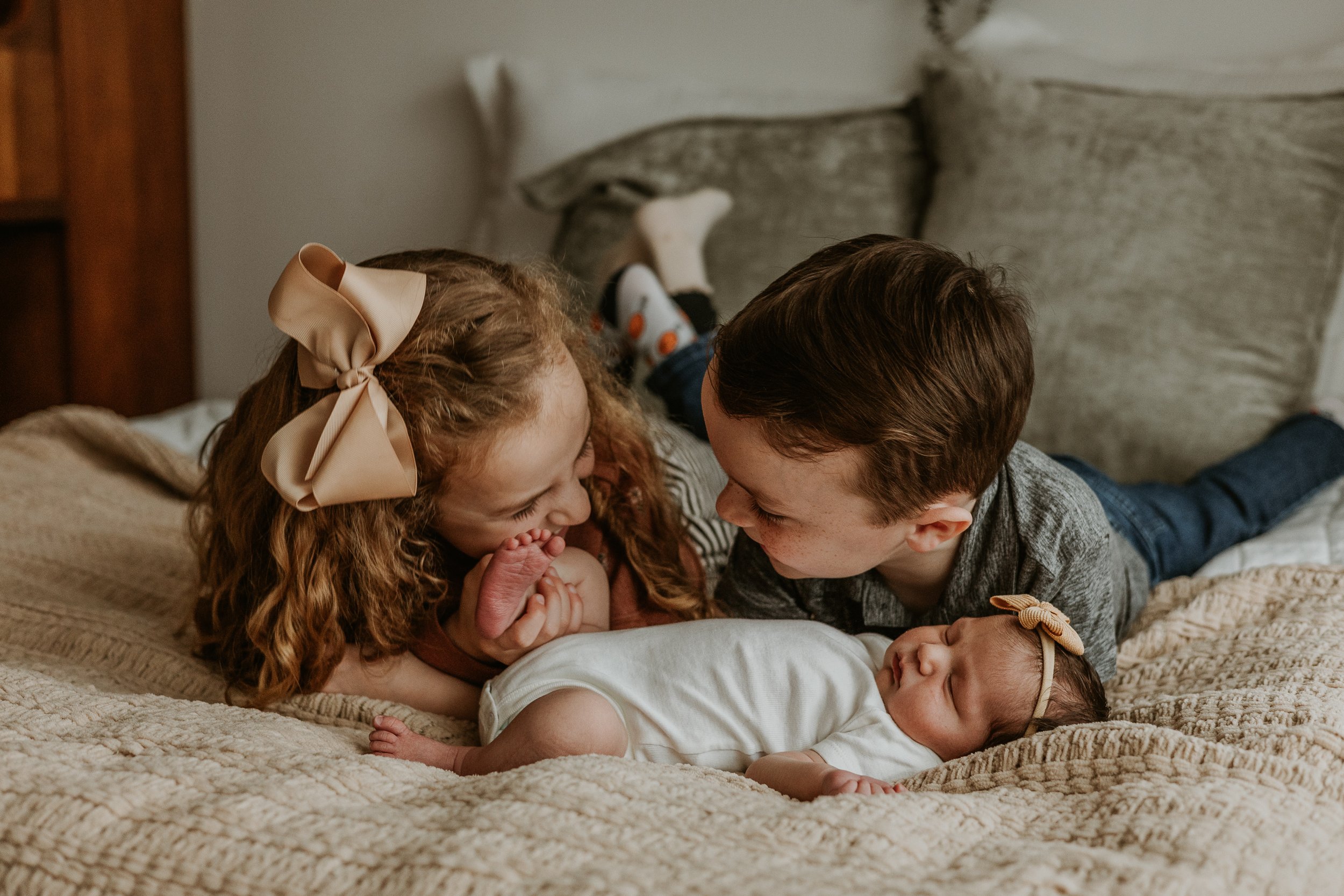 Two siblings giggle at one another as their newest sister sleeps peacefully in front of them.