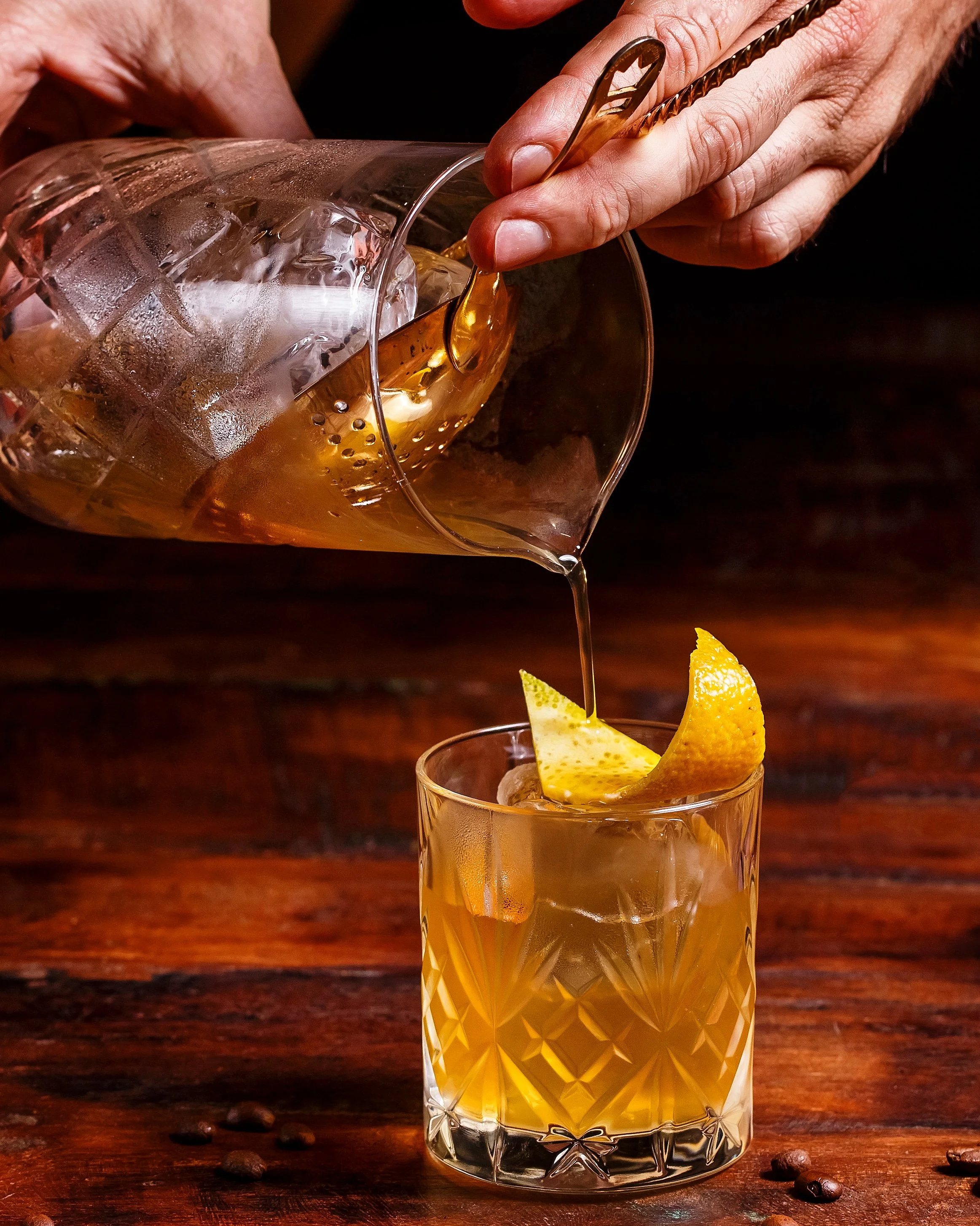 A person pouring a cocktail from a mixing glass into a glass with ice and a lemon wedge, on a wooden surface.