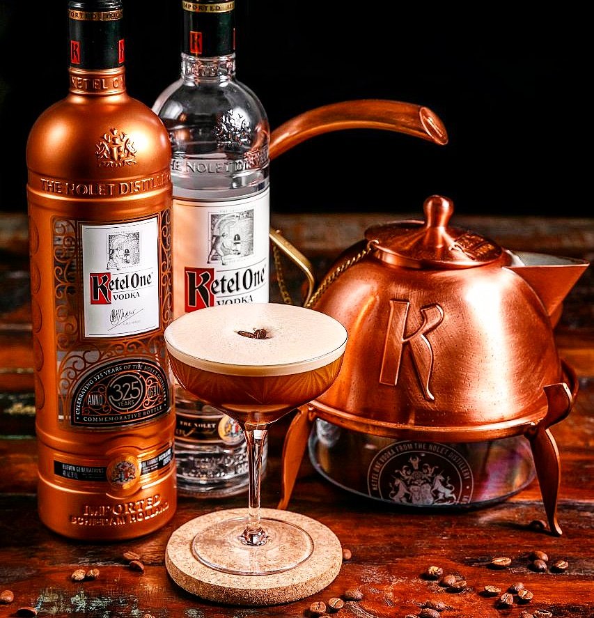 A cocktail with a frothy top and coffee beans garnish served in a coupe glass, placed on a cork coaster, alongside bottles of Ketel One vodka, a copper kettle, and a copper pot, with coffee beans scattered on a wooden surface.