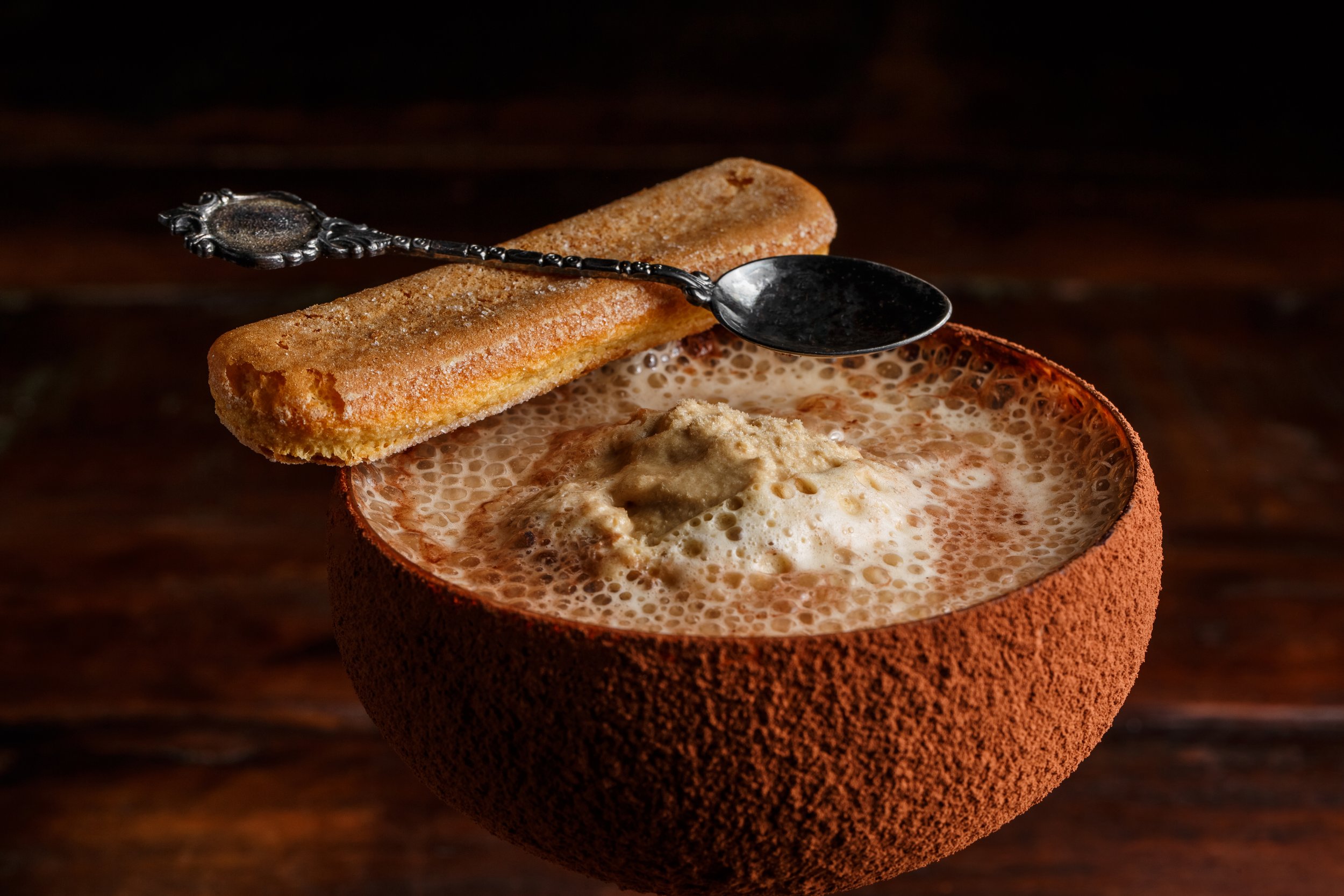 A bowl of hot chocolate topped with whipped cream, served in a clay cup with a sugar-dusted churro on top and a decorative silver spoon resting on the churro.