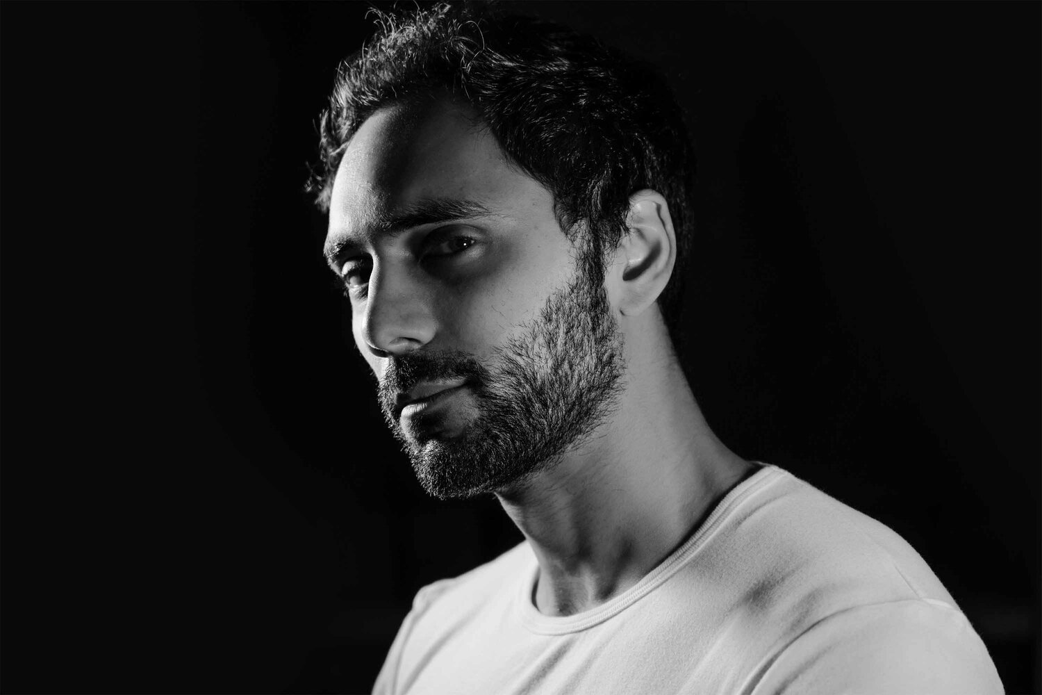 Black and white close-up portrait of a man with dark hair and beard, looking serious, with dramatic lighting against a dark background.