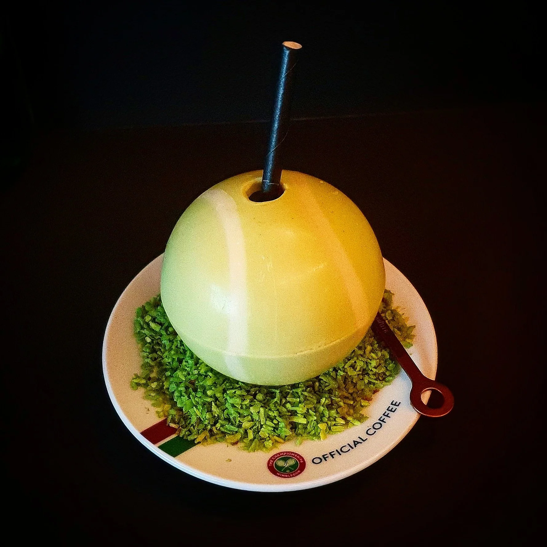 A white chocolate ball with a hole and a blue candle inside, sitting on a plate with green decorative grass and a small brown spoon, on a dark surface.
