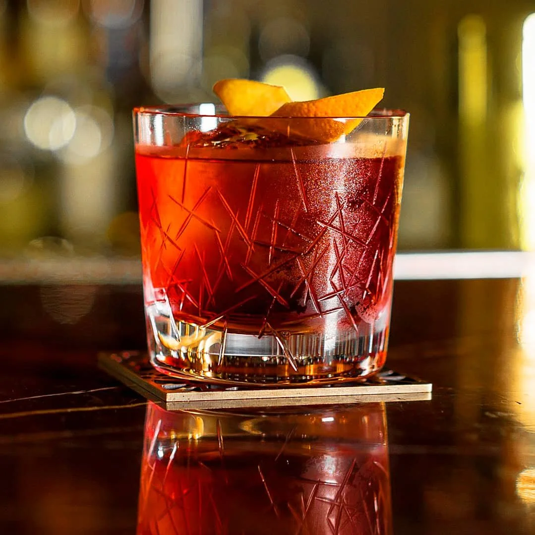 A glass of red cocktail with ice, garnished with a citrus peel, on a wooden surface with blurred background.