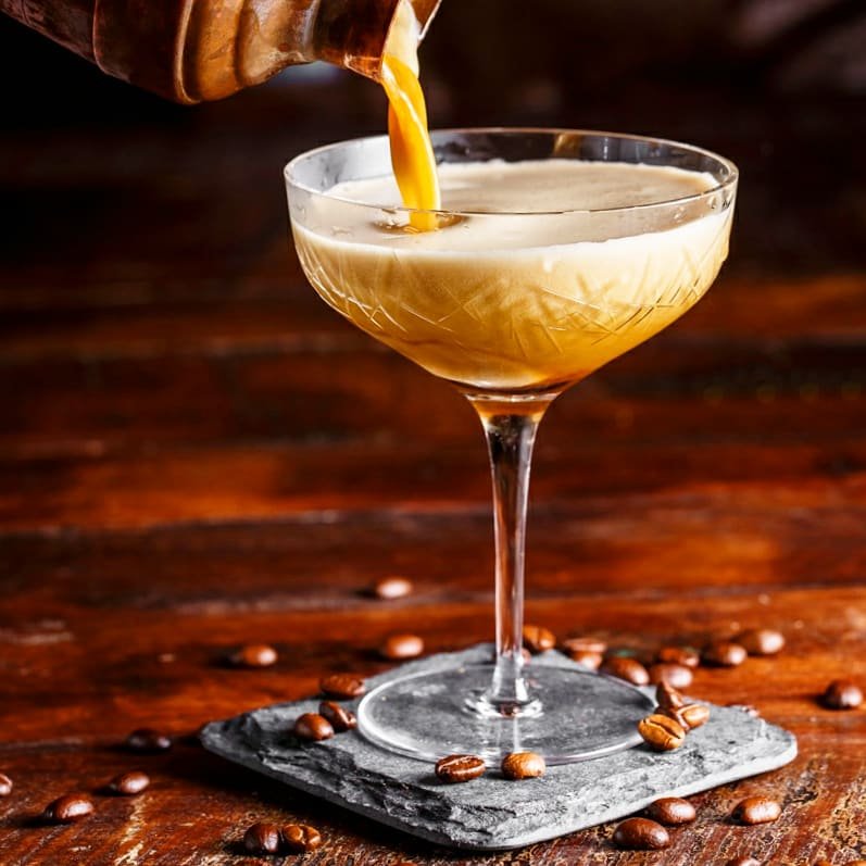 A cocktail in a coupe glass being filled with a light-colored drink over a wooden surface with scattered coffee beans.
