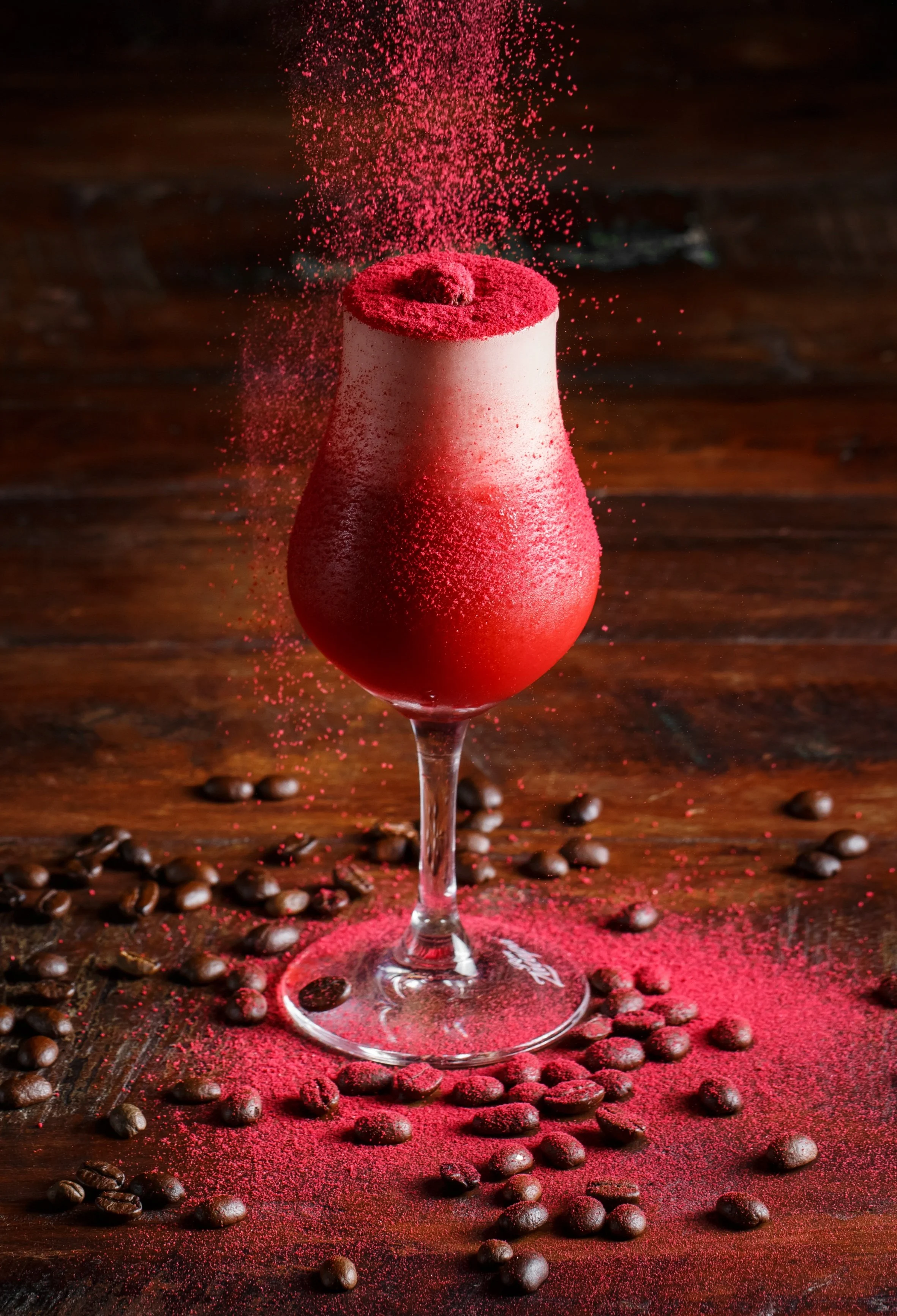 A red cocktail drink in a tall, curved glass on a wooden surface, surrounded by scattered coffee beans and red powder, with additional red powder falling into the drink.