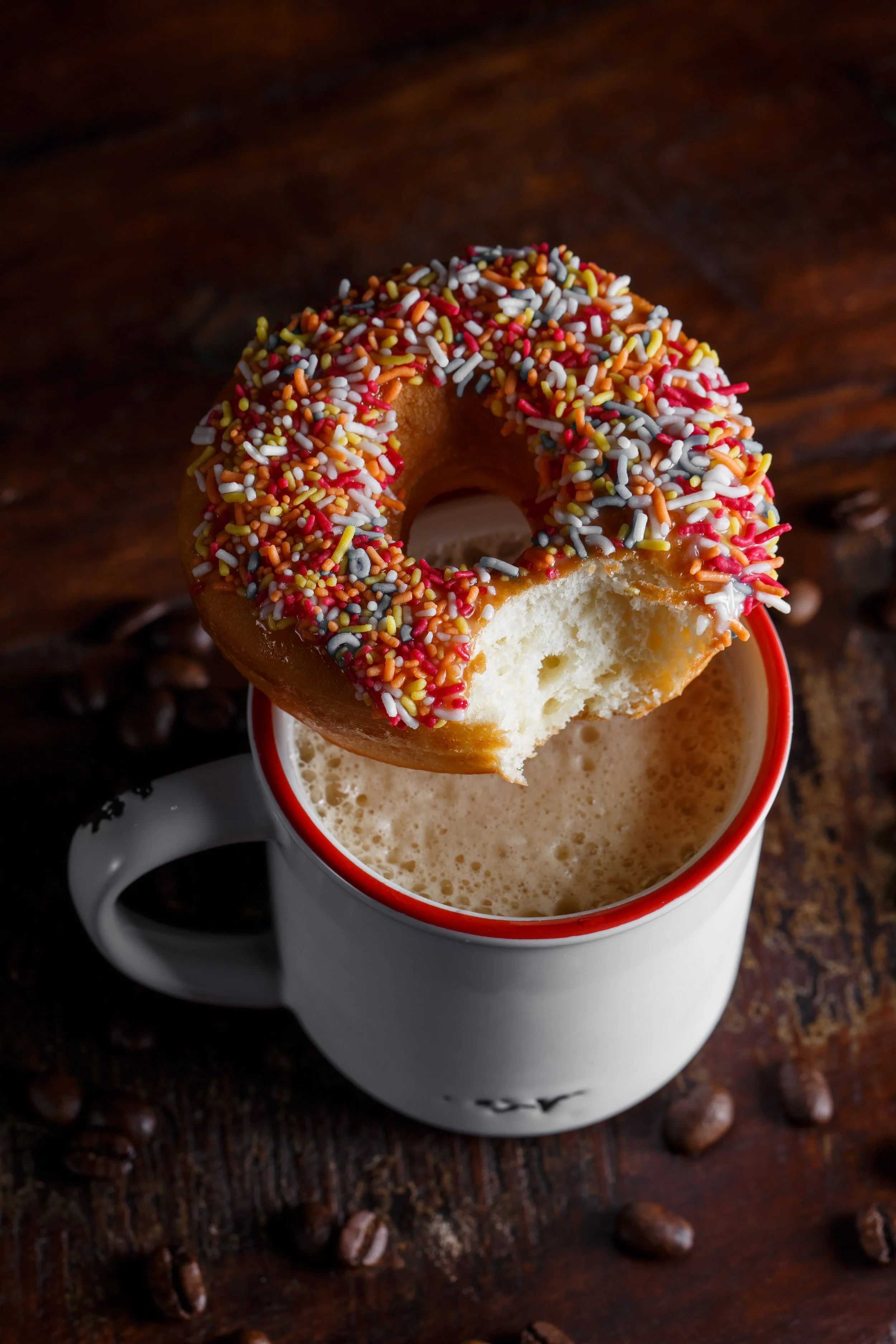 A donut with colorful sprinkles resting on the rim of a mug filled with a frothy beverage on a wooden surface.