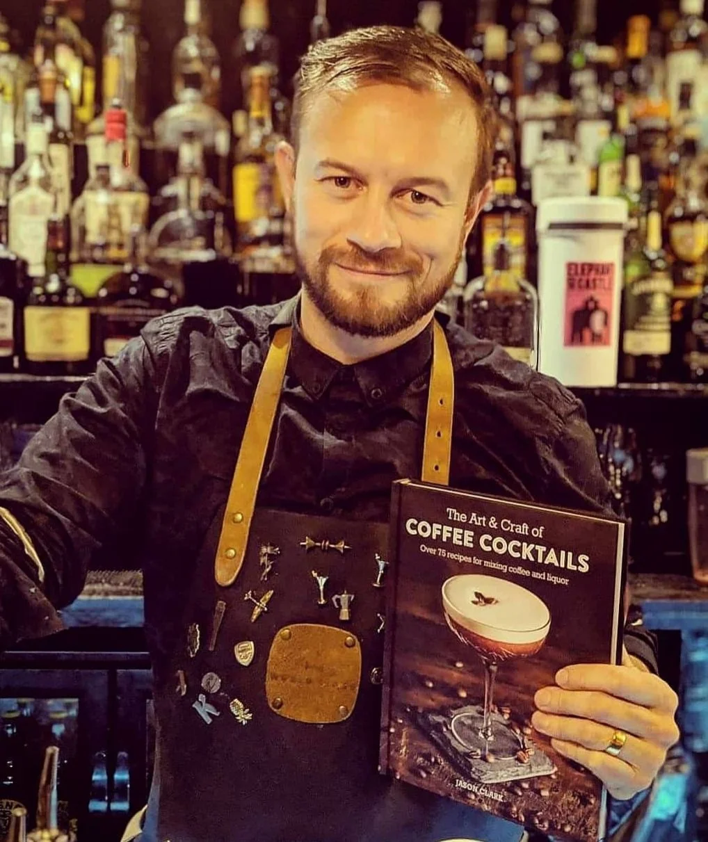 A man with a beard and short hair, wearing a dark shirt and a brown apron with pins, standing behind a bar and holding a book titled 'The Art & Craft of Coffee Cocktails' in front of a background filled with liquor bottles.