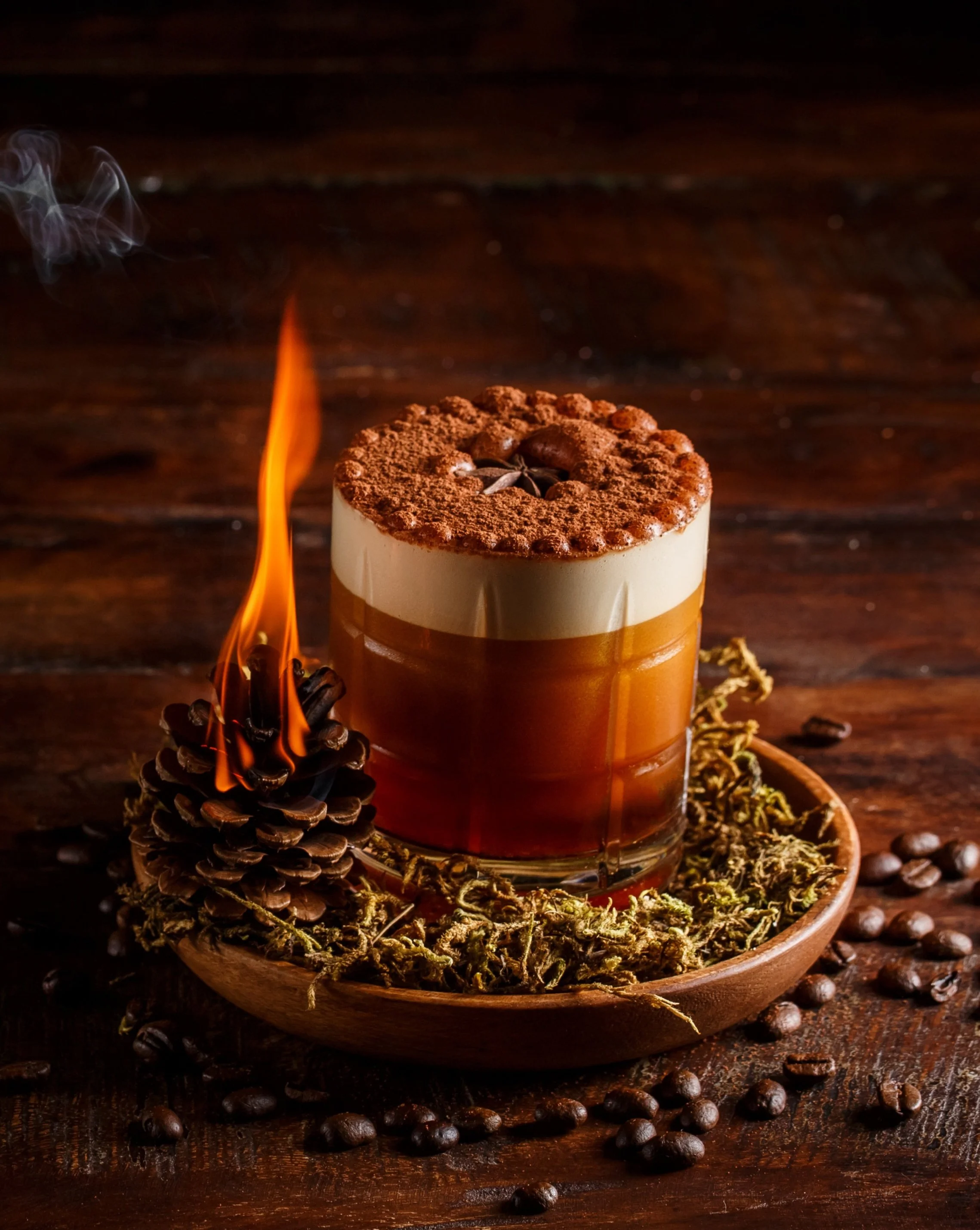 Layered coffee drink with foam topping in a glass, surrounded by coffee beans, a pine cone, and moss on a wooden tray, with a small flame nearby.