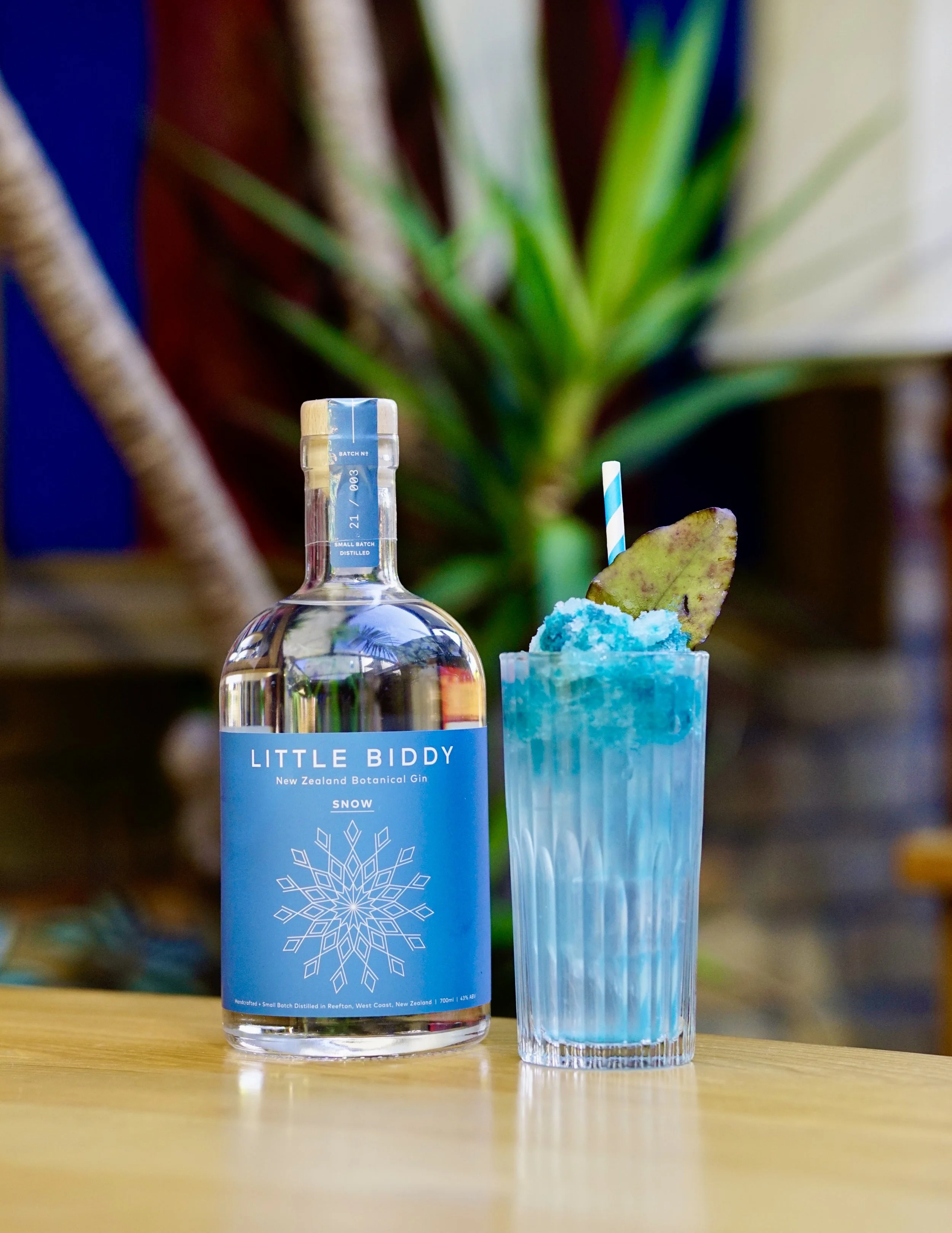A bottle of Little Biddy New Zealand Botanical Gin labeled Snow next to a blue frozen cocktail garnished with a leaf and a blue and white striped straw on a wooden table with plants in the background.