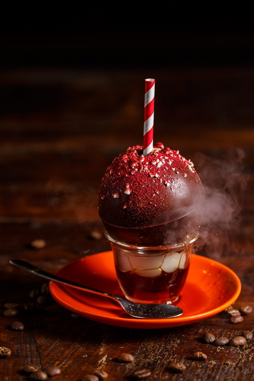 A dessert in a small glass cup with layers of red syrup and white jelly, topped with a red and white candy cane striped straw, red sprinkle topping, and small white sugar crystals, served on an orange saucer with a spoon on a wooden table scattered w