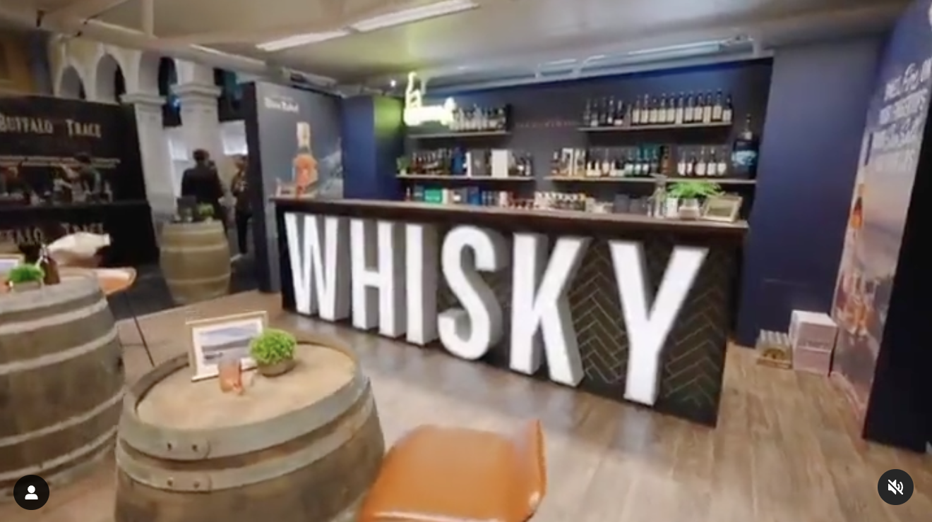 Bar area with large illuminated letters spelling 'WHISKY'. The bar has shelves stocked with bottles, and there are tables made from barrels with small plants and menu cards on them.