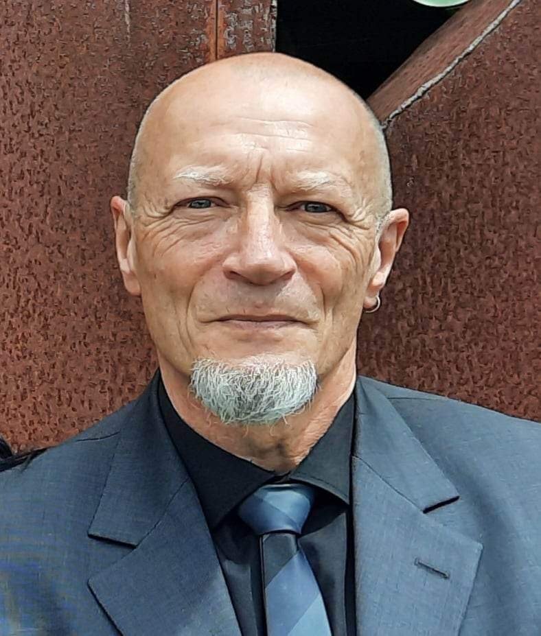 A portrait of an elderly man with a bald head, a white goatee, blue eyes, and a small earring in his left ear. He is wearing a dark suit and a blue striped tie, standing against a brick wall.