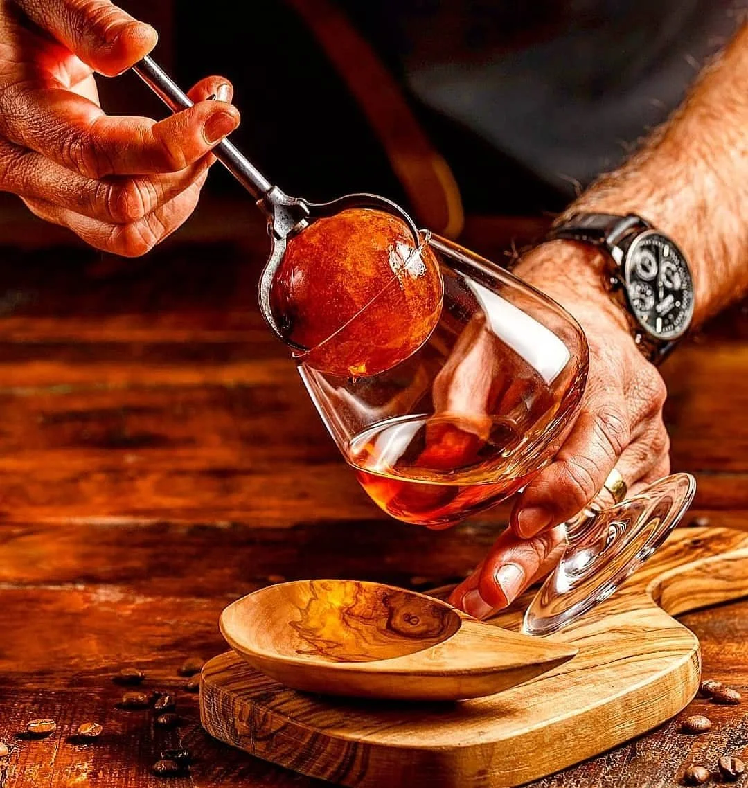 Person pouring whiskey from a decanter into a glass with an ice sphere using tongs, on a wooden table with coffee beans and a small wooden spoon.