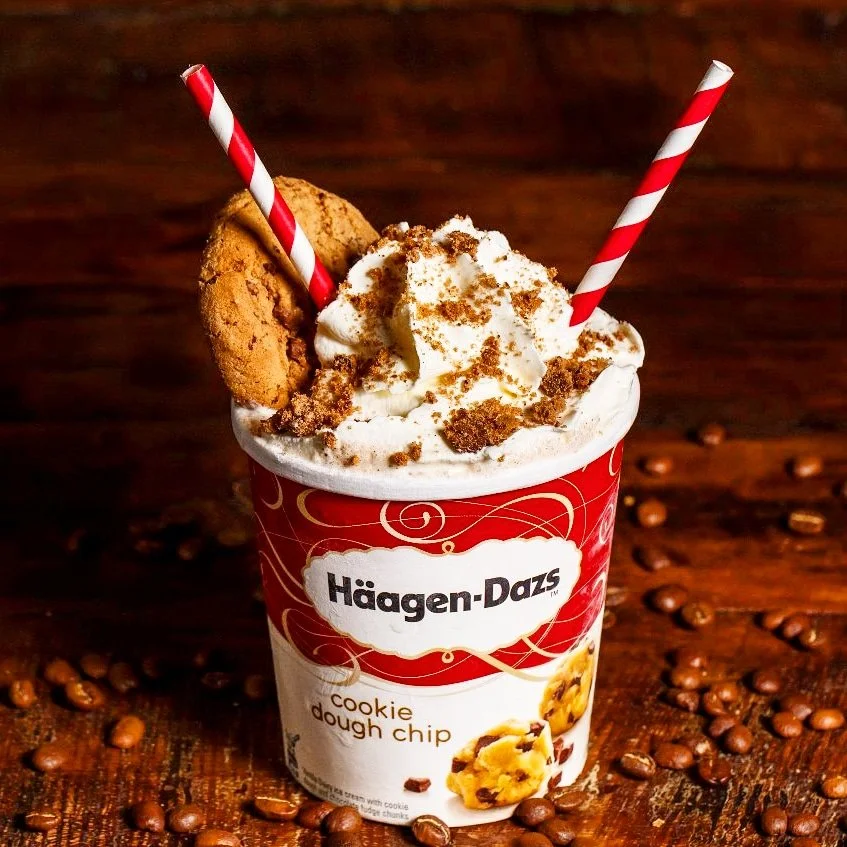 A cup of Häagen-Dazs cookie dough ice cream topped with whipped cream, cookie dough chunks, and crumbled cookies, with two red and white striped straws, surrounded by coffee beans on a wooden surface.