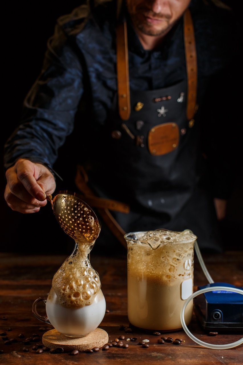 Man preparing a layered coffee drink, with foam spilling over from a small glass vessel into a cup, on a wooden surface surrounded by coffee beans.