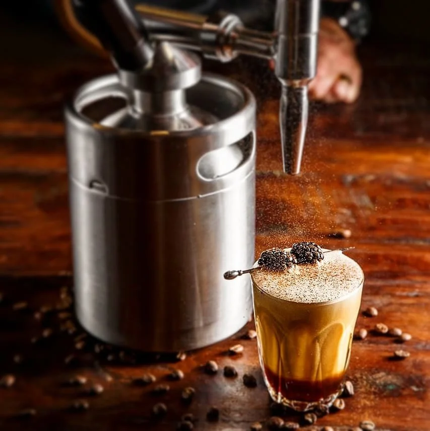 An espresso machine dispensing coffee into a glass with foam and garnish, surrounded by scattered coffee beans on a wooden surface.
