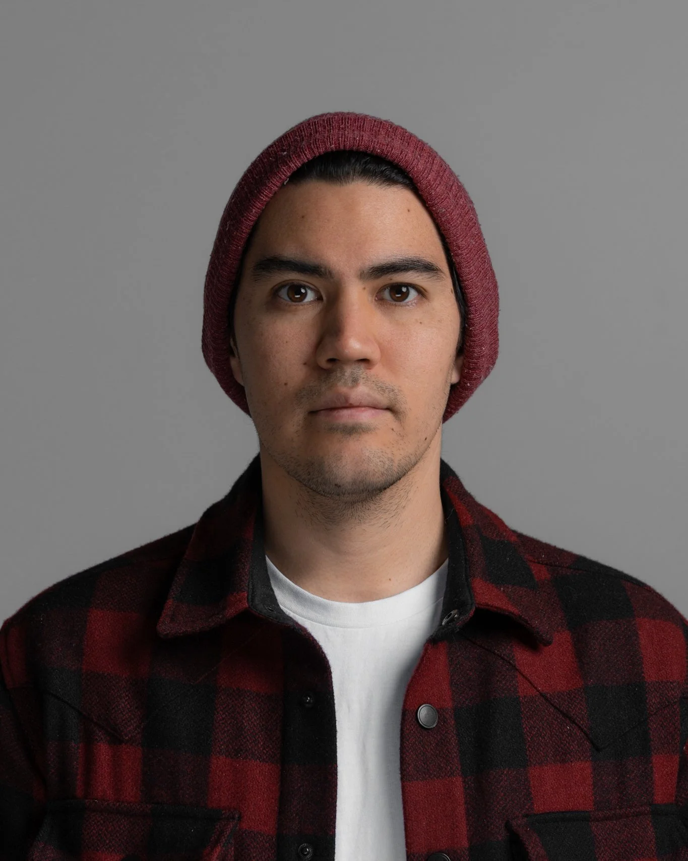 Portrait of a young man wearing a red knit beanie, a black and red plaid shirt, and a white T-shirt against a plain gray background.