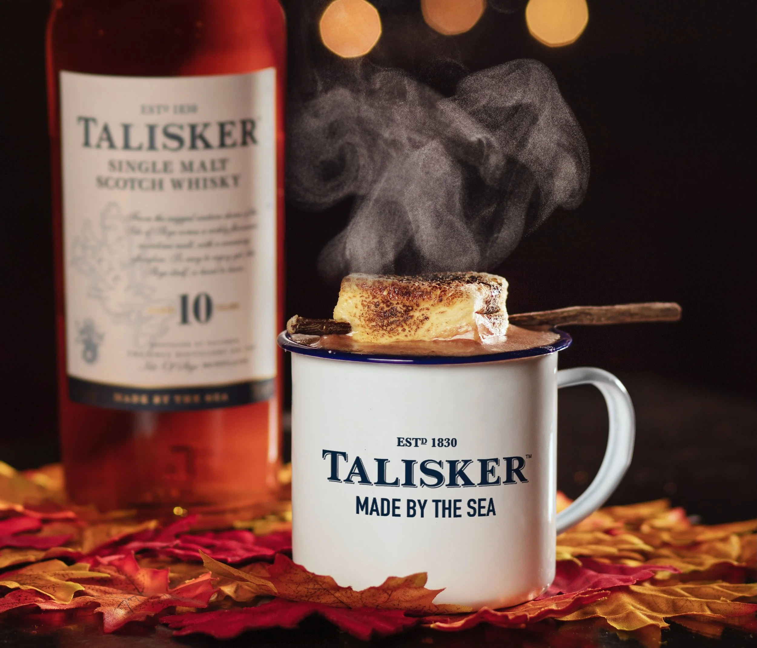 A steaming cup of hot beverage with a piece of bread on a skewer resting on top, placed on an autumn-themed table with fallen leaves, with a bottle of Talisker single malt Scotch whisky in the background.