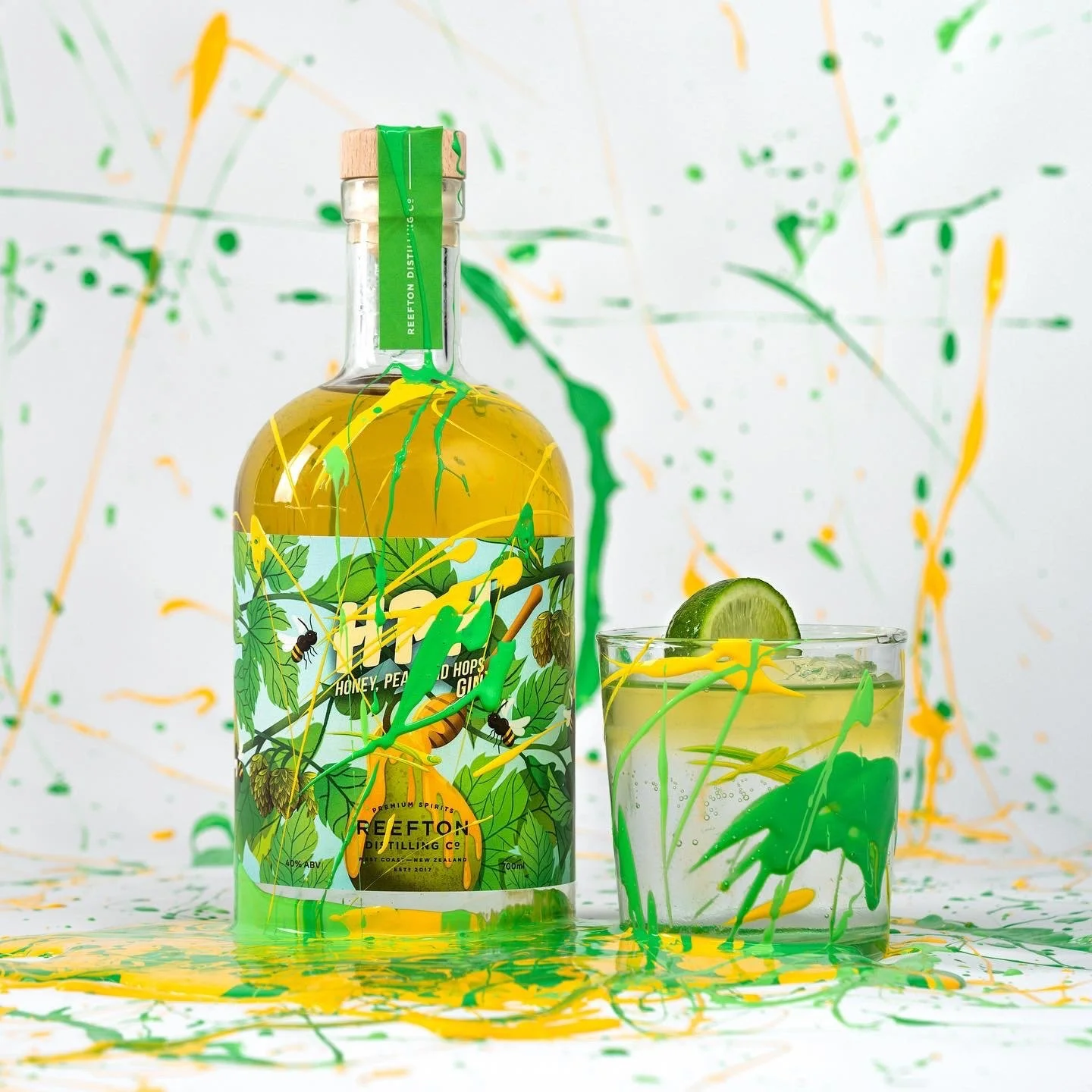 A bottle of Reefton spirit with a label showing honey, peach, hops, and ginger, alongside a glass with a green lime wedge, all splattered with yellow, green, and orange paint.