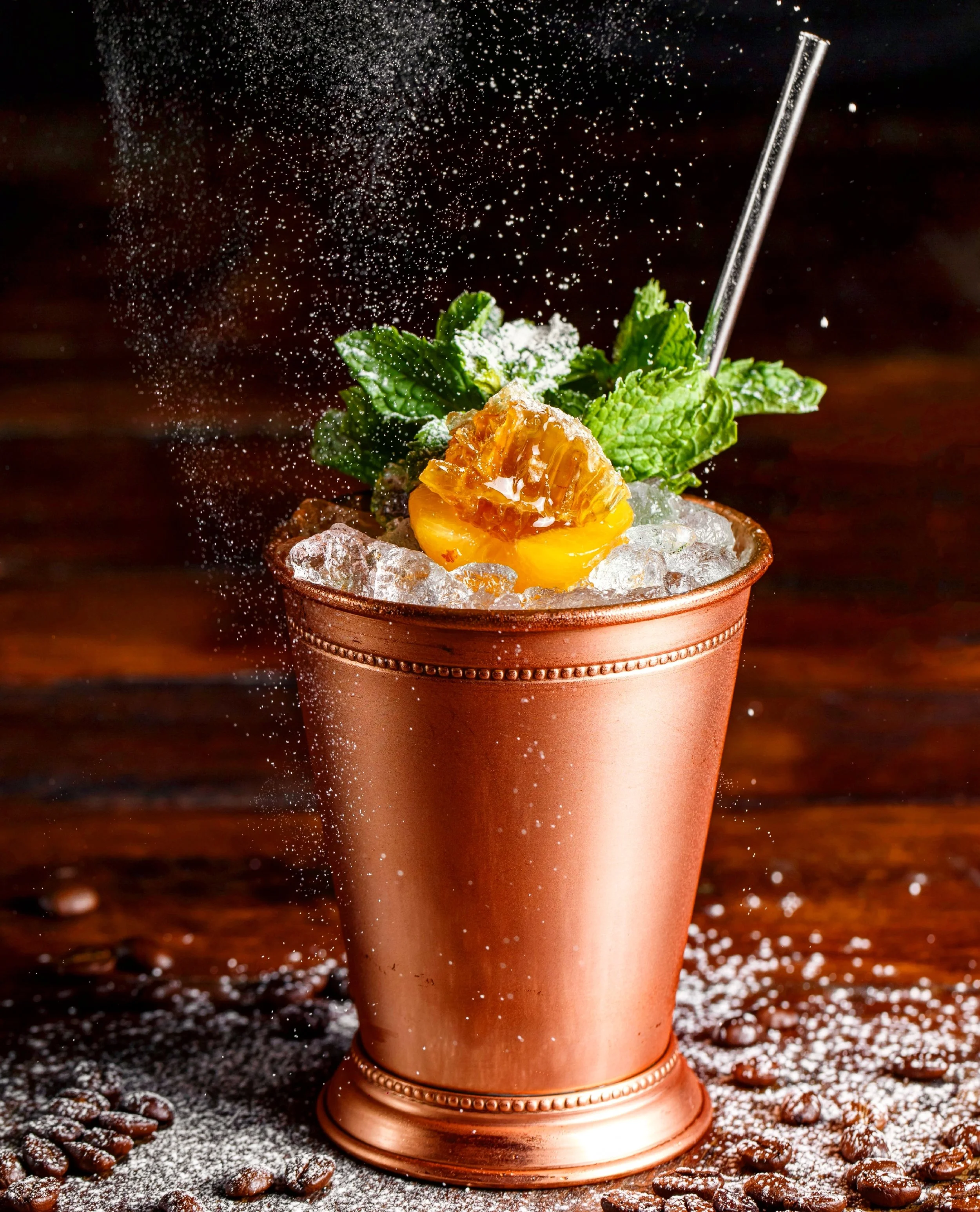 A copper cup filled with ice, garnished with a yellow half peach, fresh mint leaves, and a metal straw, with sugar and coffee beans scattered on a wooden surface.