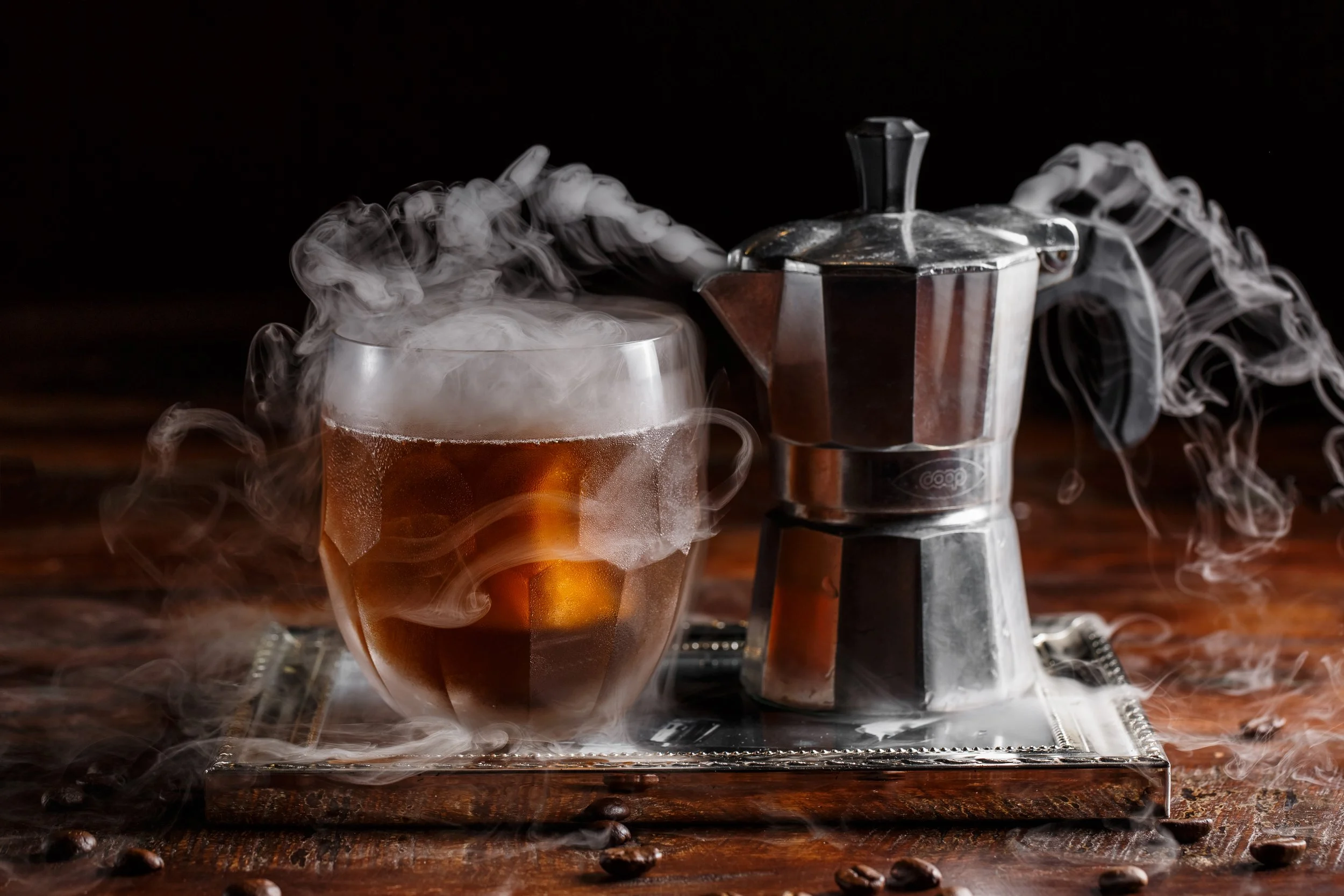 A steaming cup of coffee and a moka pot on a silver tray with coffee beans scattered, on a wooden surface.