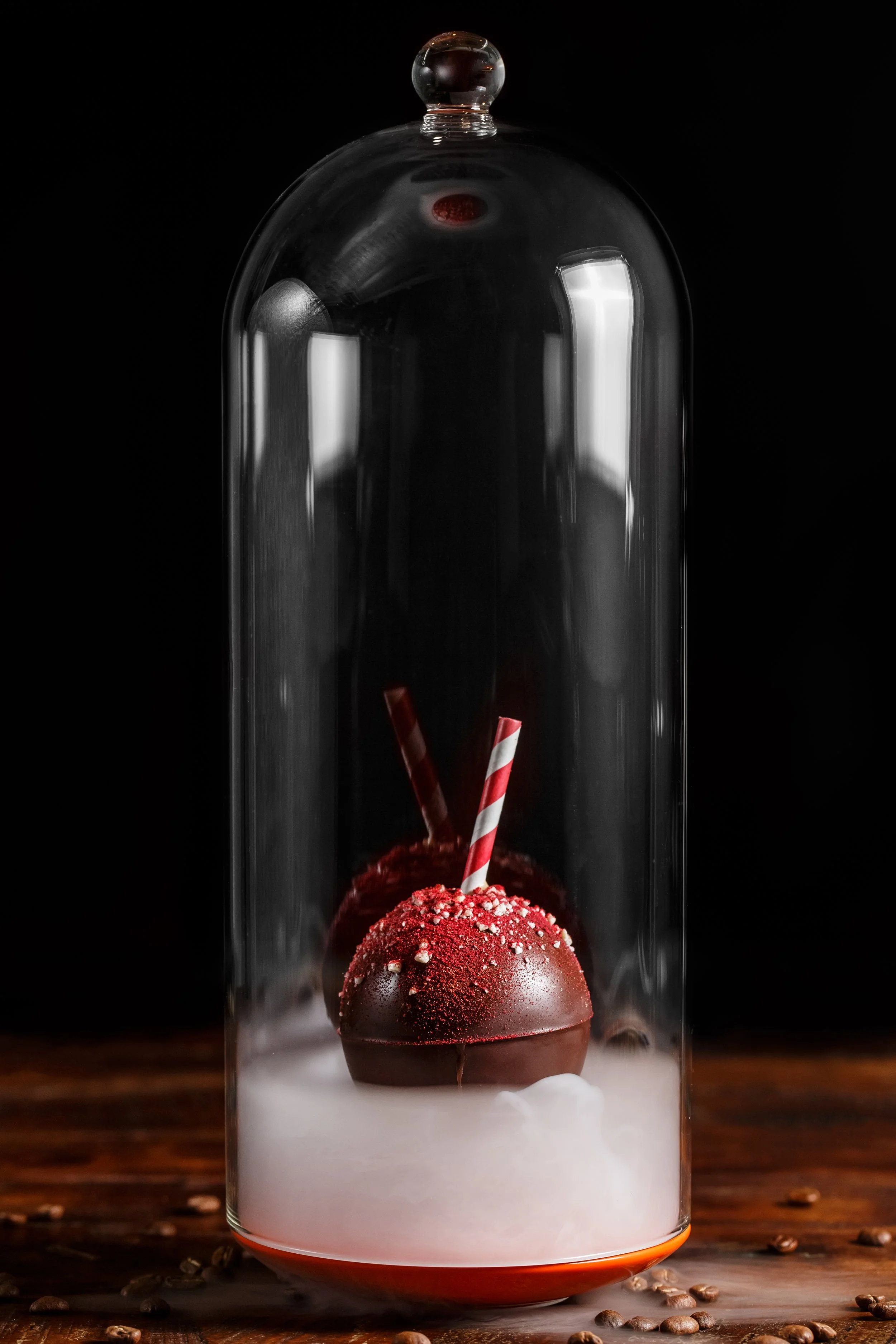 A glass dome encases two red chocolate candies topped with crushed candies and two striped straws, sitting on a wooden surface with scattered coffee beans.