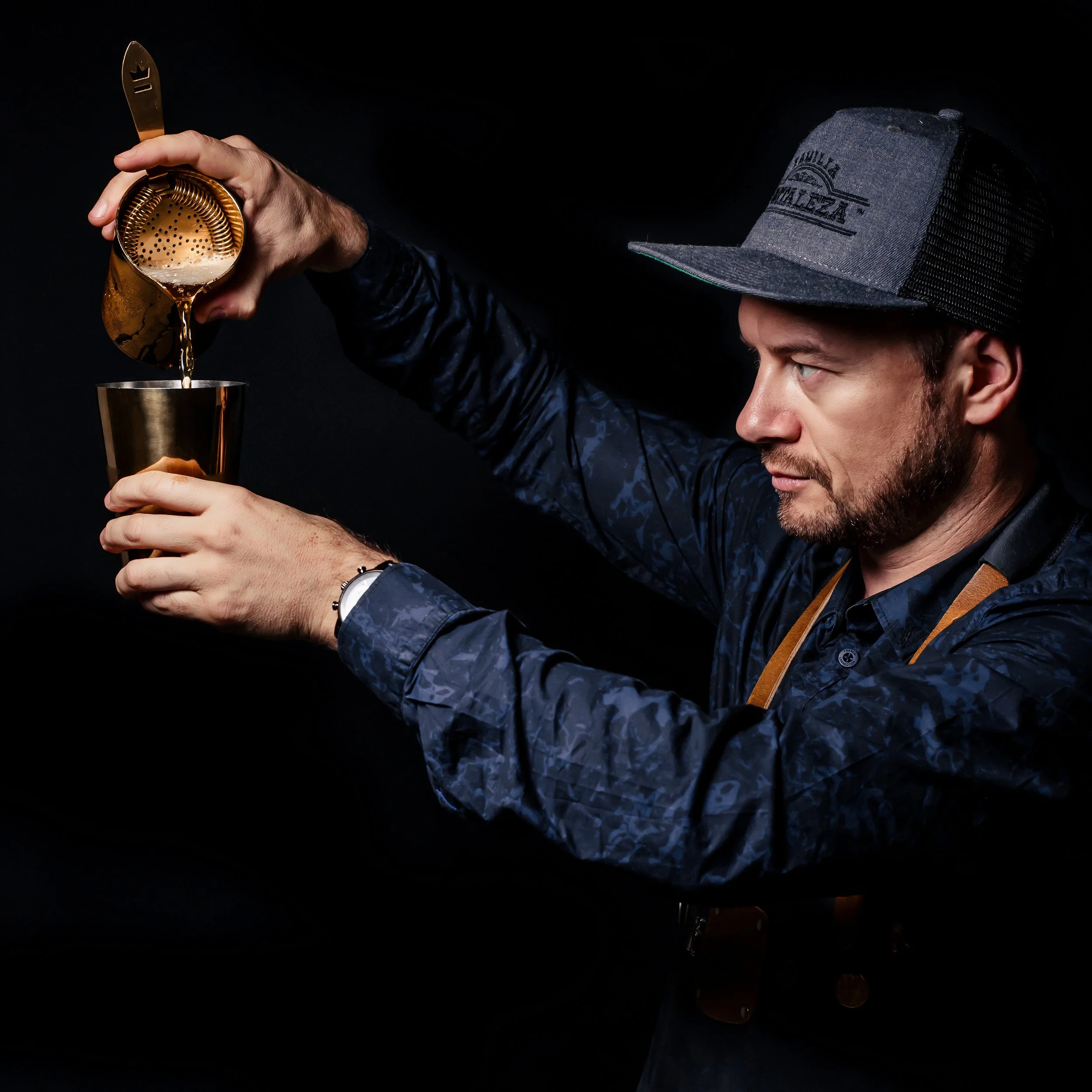 A man wearing a gray cap and a dark patterned shirt is pouring a drink from a metallic cocktail shaker into a metallic cup, with a dark background.