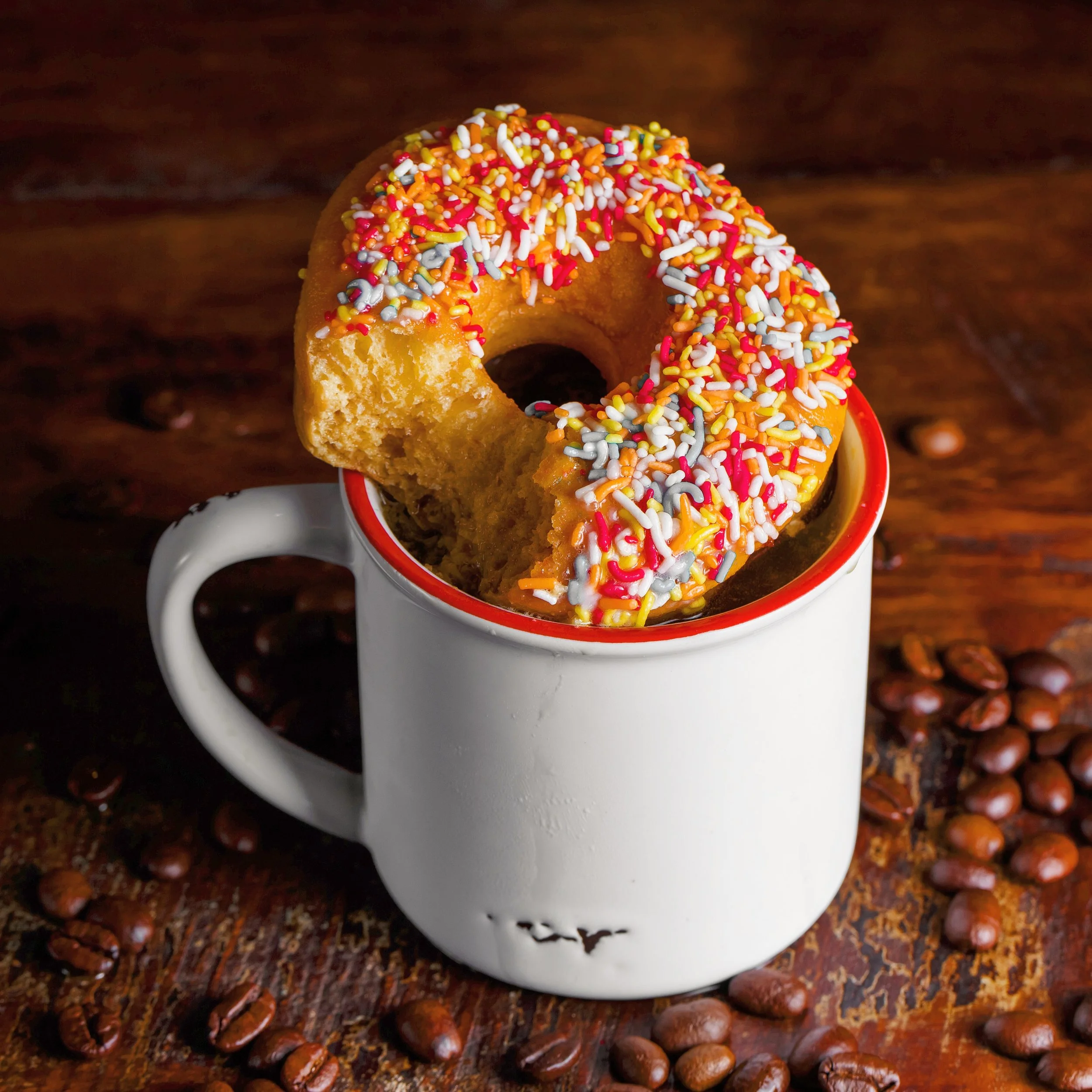 A coffee mug with a partially eaten donut topped with colorful sprinkles on top of a wooden surface surrounded by scattered coffee beans.