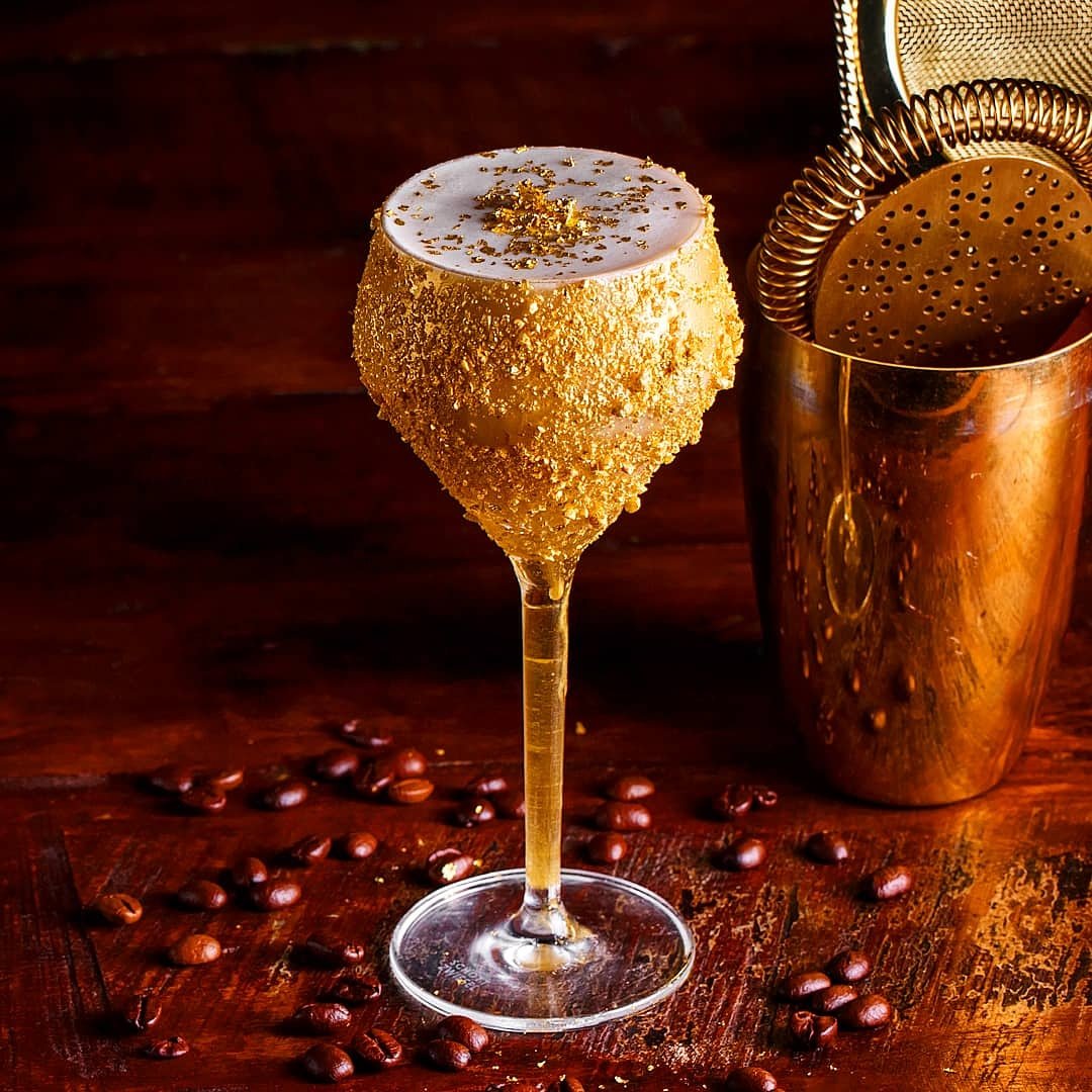 A fancy cocktail in a tall glass with a sugar and gold coating, garnished with gold flakes on top, placed on a wooden surface surrounded by coffee beans, with a cocktail shaker in the background.