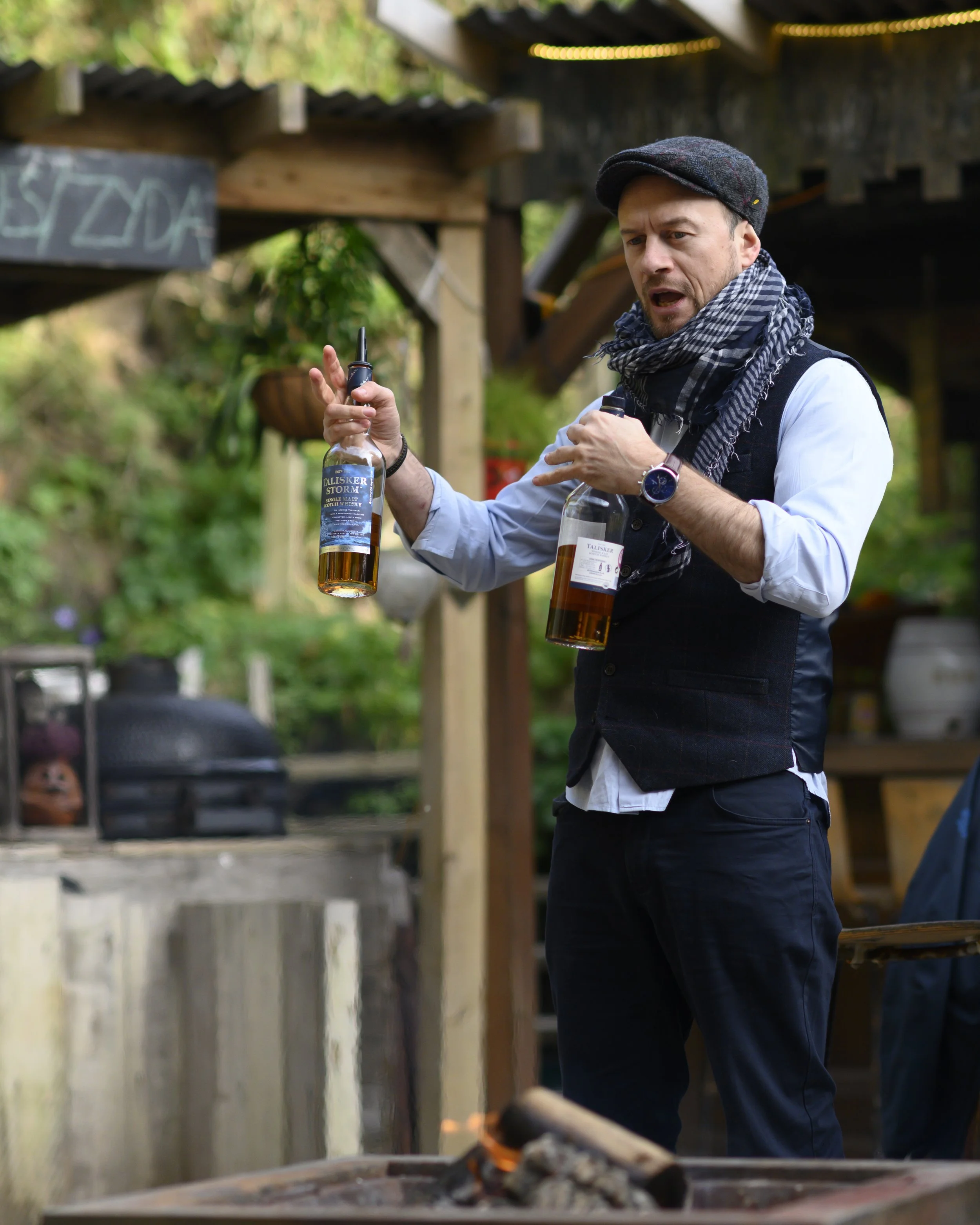 A man in a black vest, light blue shirt, and scarf is holding two bottles of whiskey and talking outdoors near a fire pit.