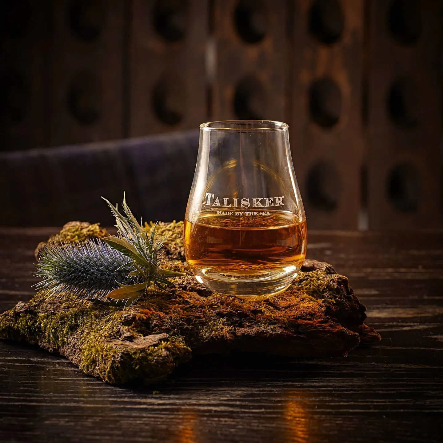 A glass of whisky on a piece of mossy wood with a sprig of lavender, set on a dark wooden table against a dark background.