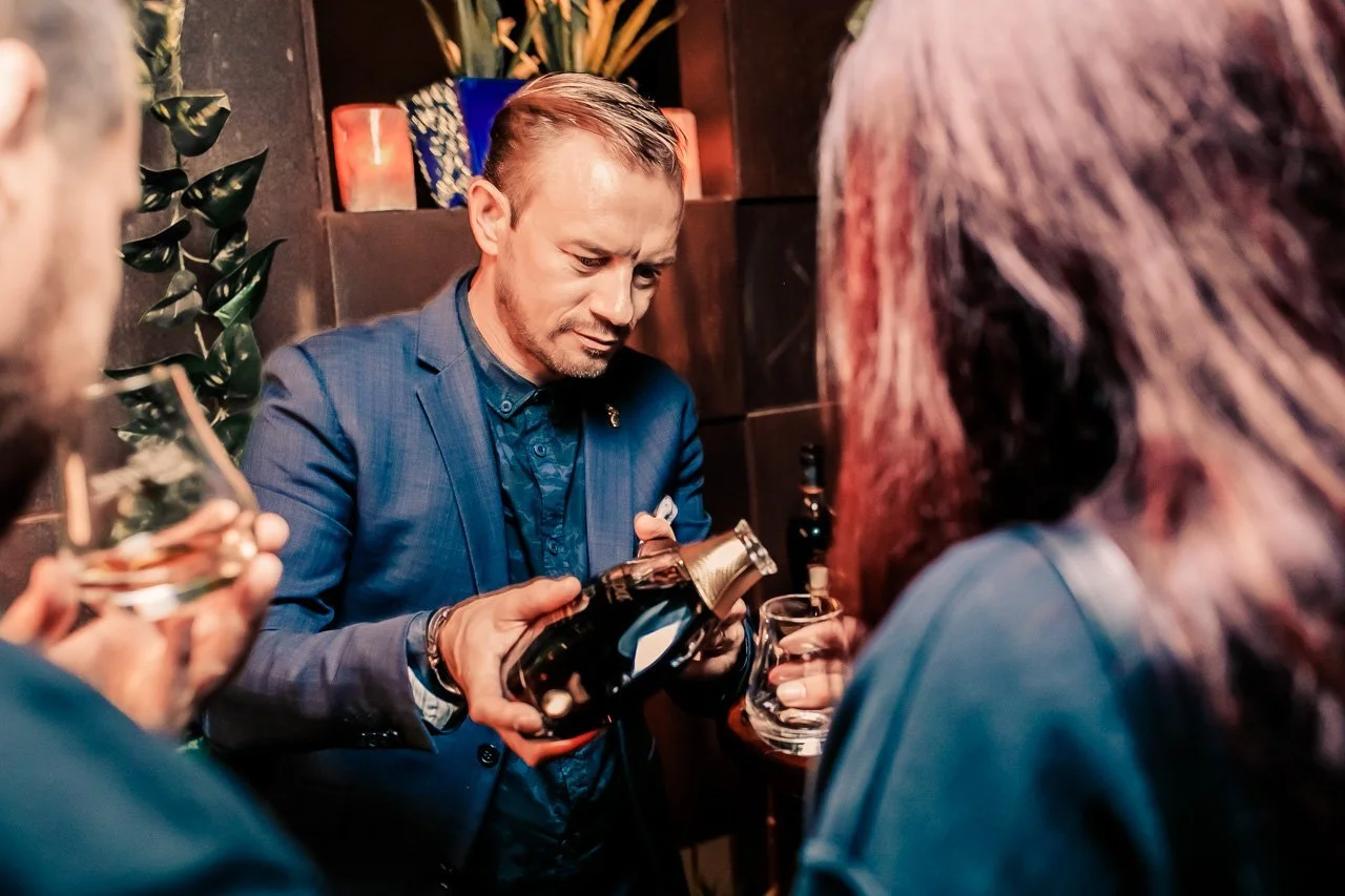 A man in a blue suit pours a drink into a glass while surrounded by people in a dimly lit bar or lounge.