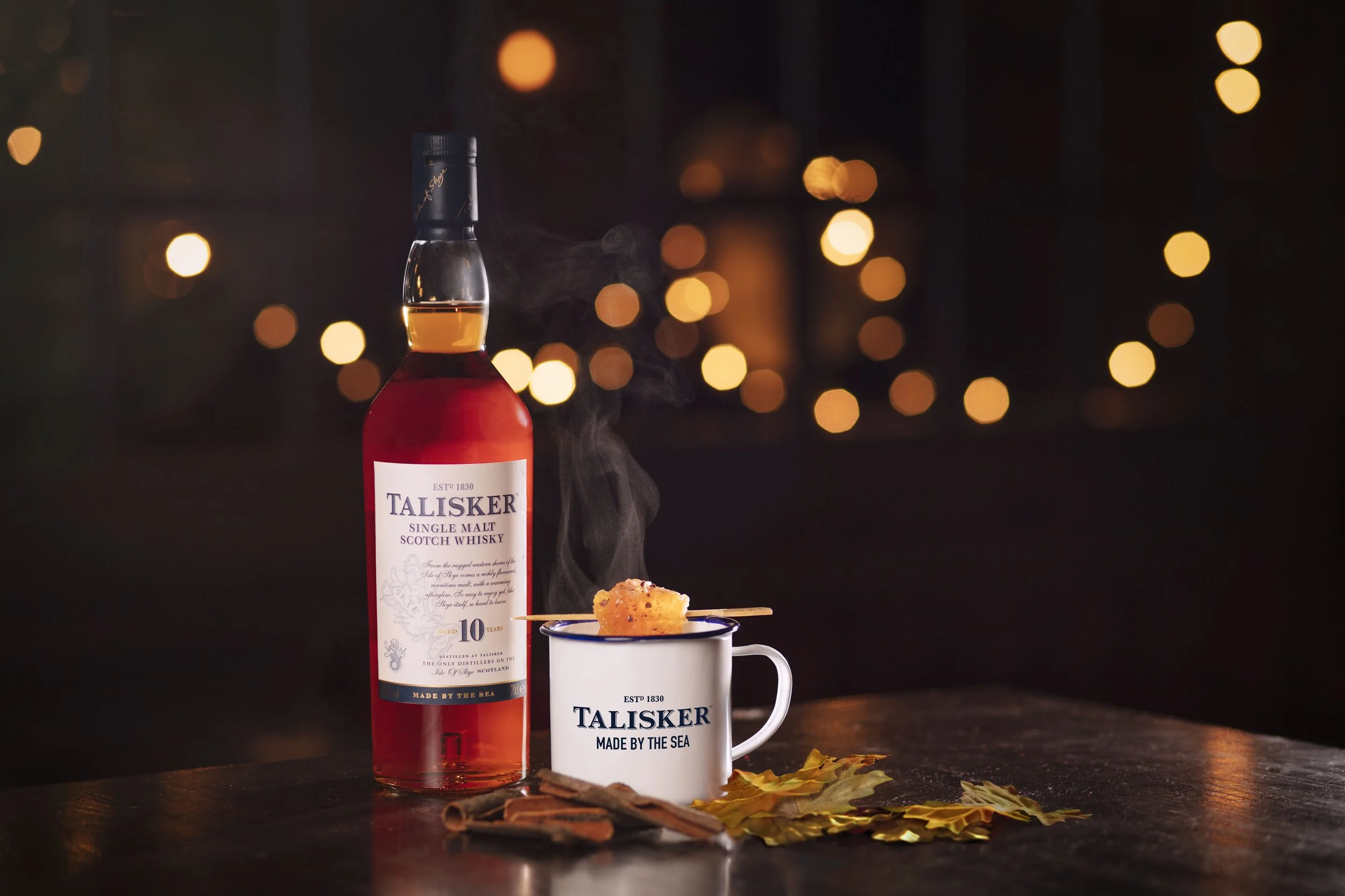 A bottle of Talisker single malt scotch whisky, a steaming mug with the Talisker logo, a skewer with a piece of grilled food, cinnamon sticks, and autumn leaves on a wooden table at night.